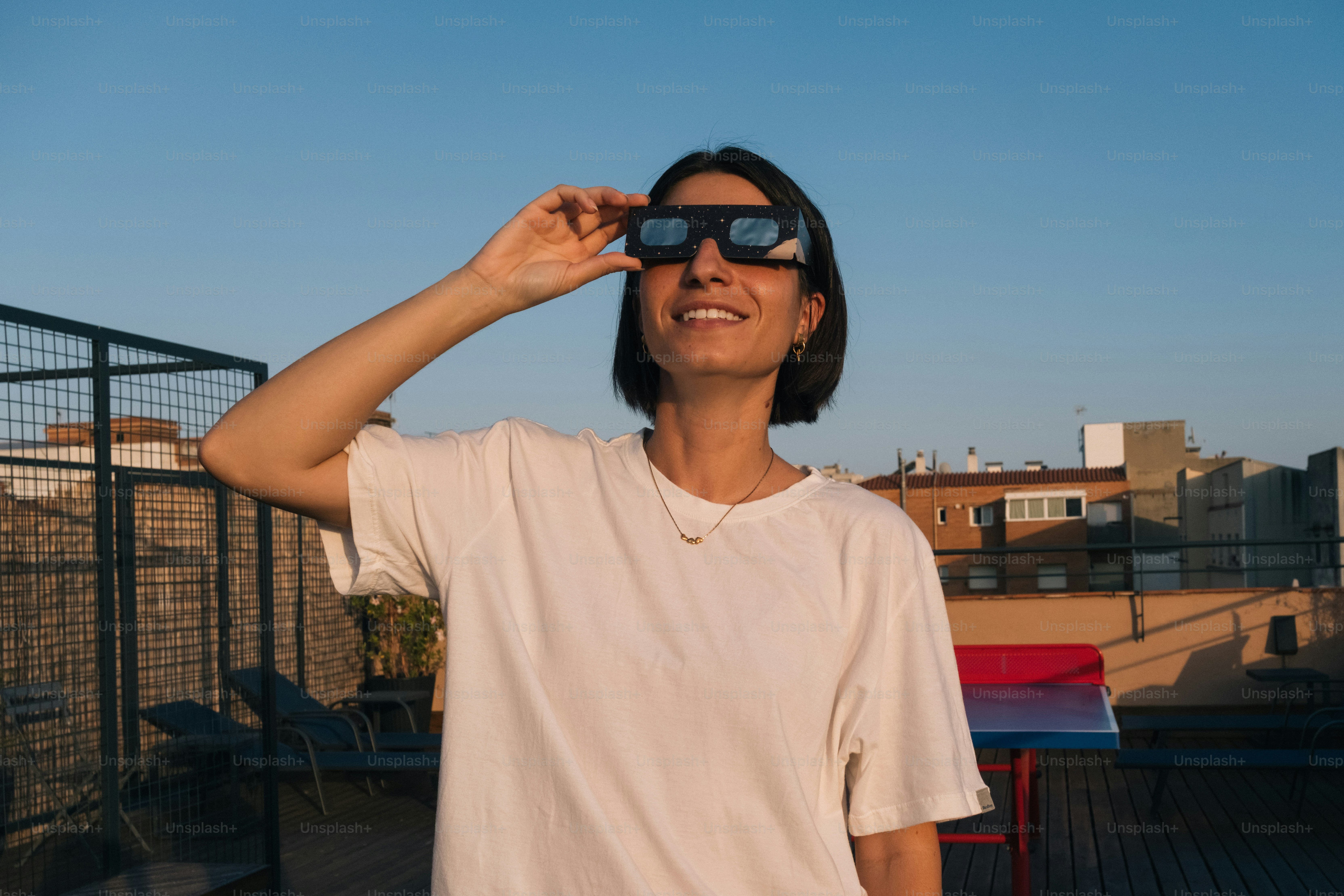 A woman wearing solar eclipse glasses on a rooftop at golden hour.