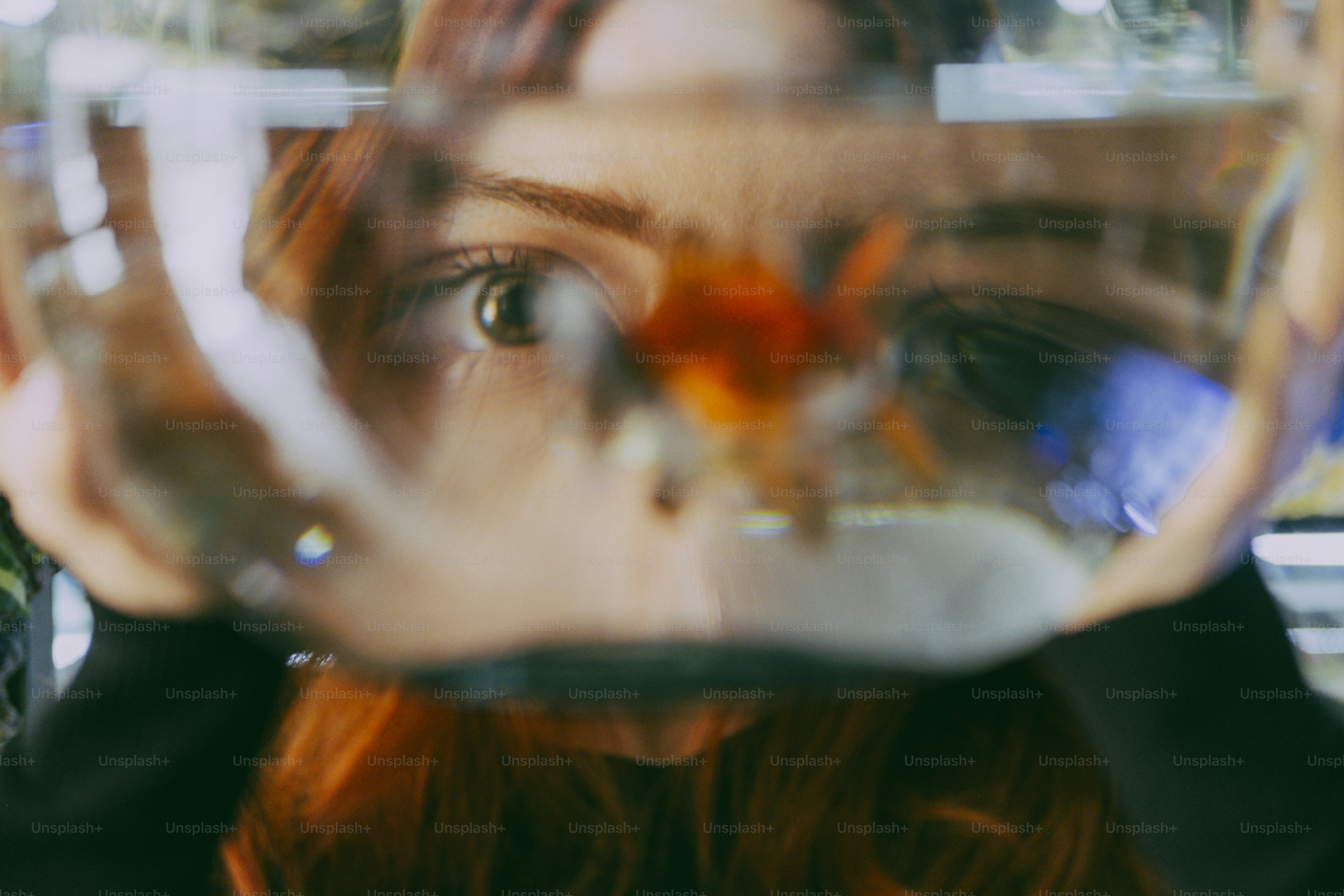 A person looking through a glass bowl containing a small orange goldfish