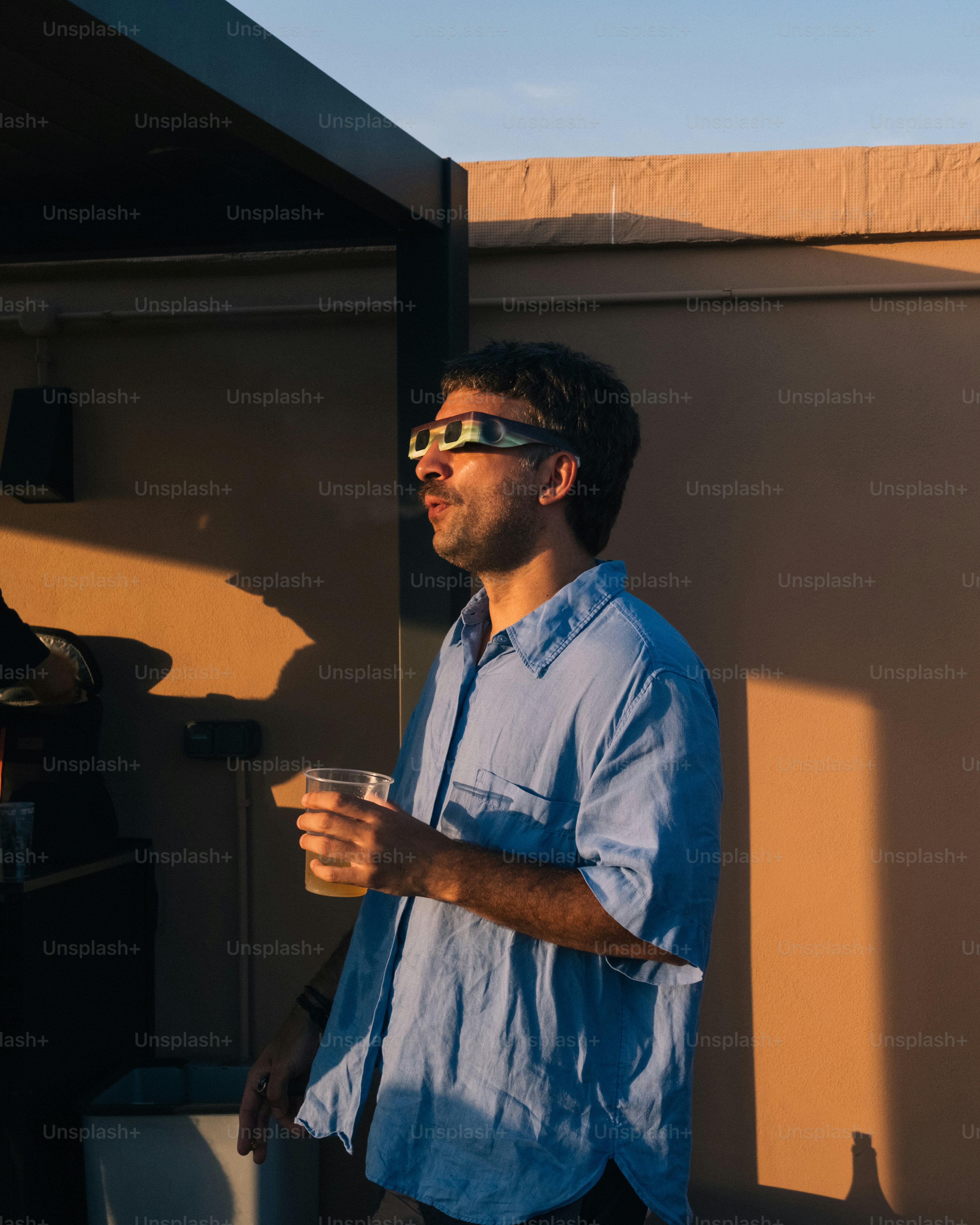 A man in a blue shirt wearing eclipse glasses and holding a plastic cup.