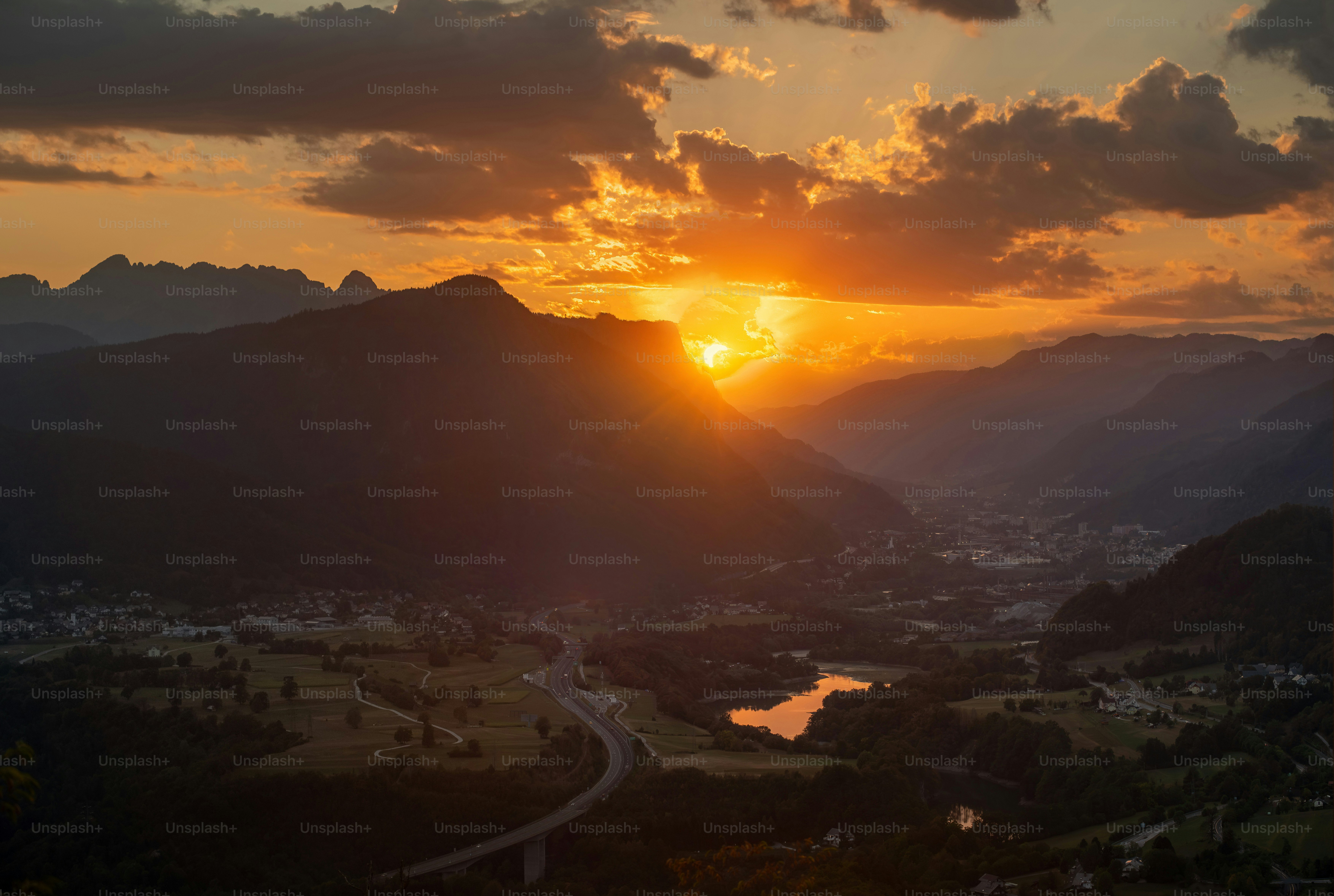 Low sun glowing behind dark mountain silhouettes above a valley with a winding road