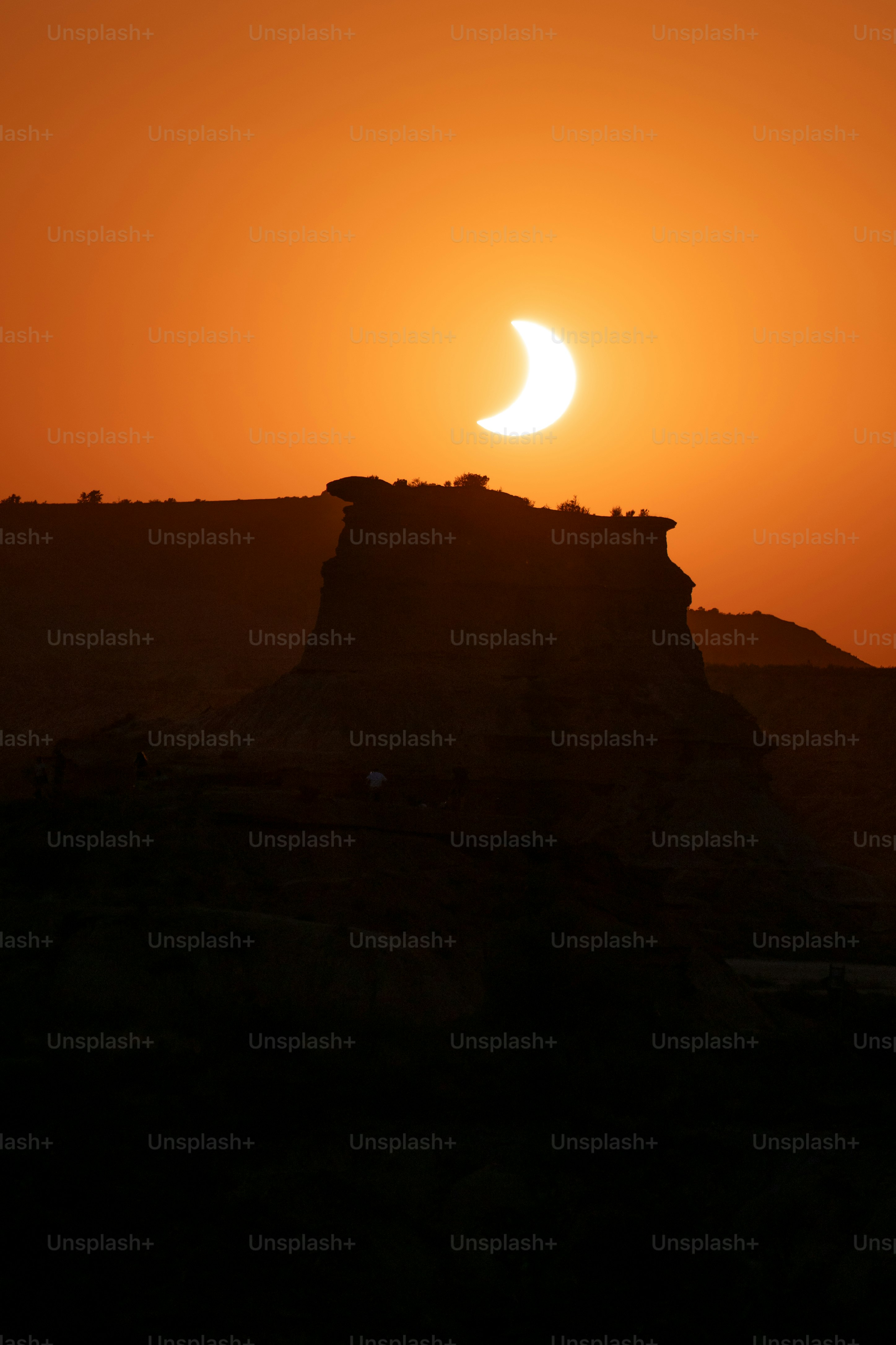 A partial solar eclipse above a dark rocky mesa in a warm orange sky