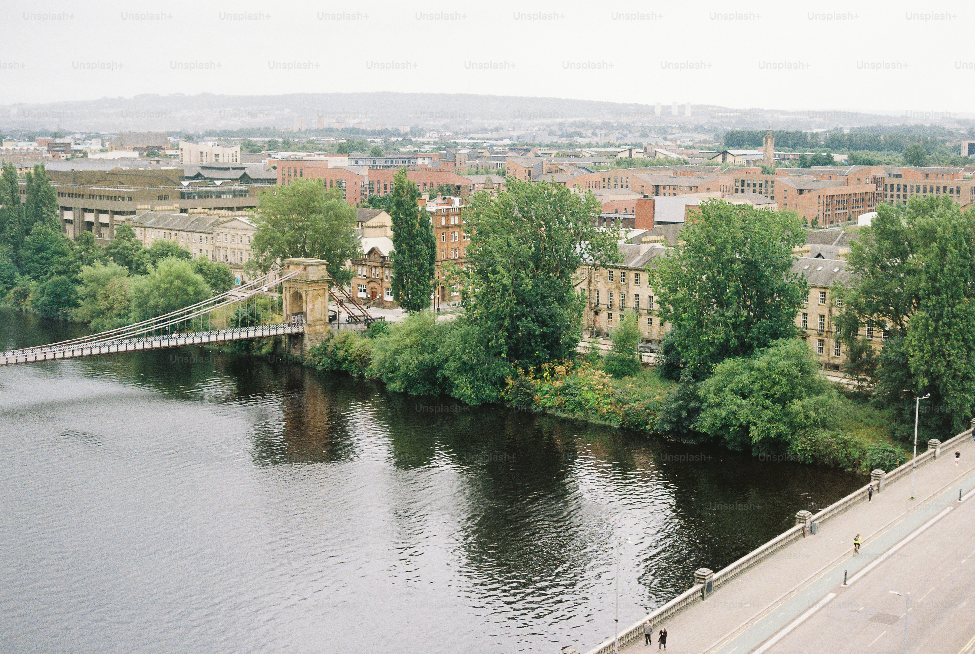 A suspension bridge over a river with buildings and trees in the background