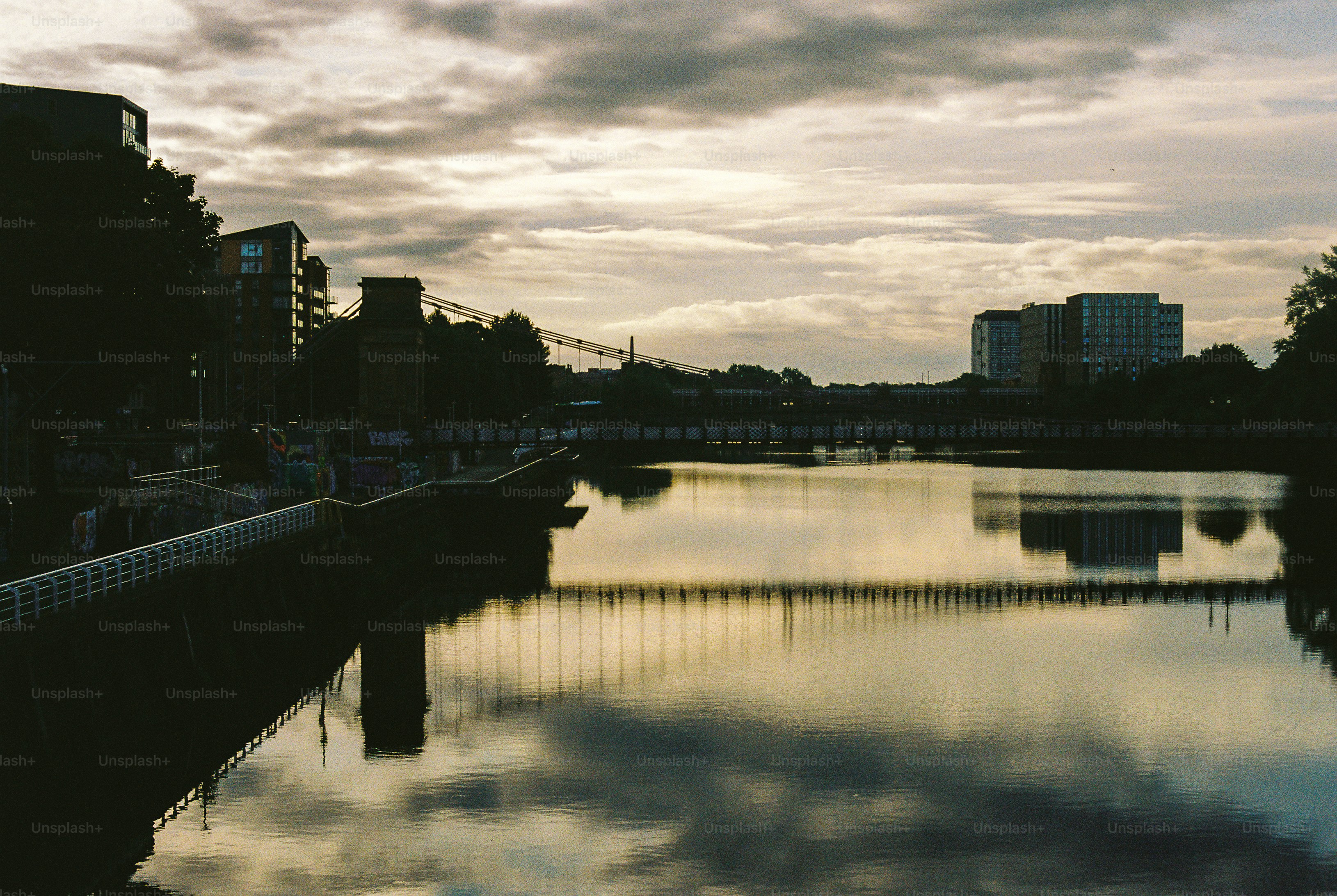 A suspension bridge over a calm river reflecting a cloudy sky at dusk