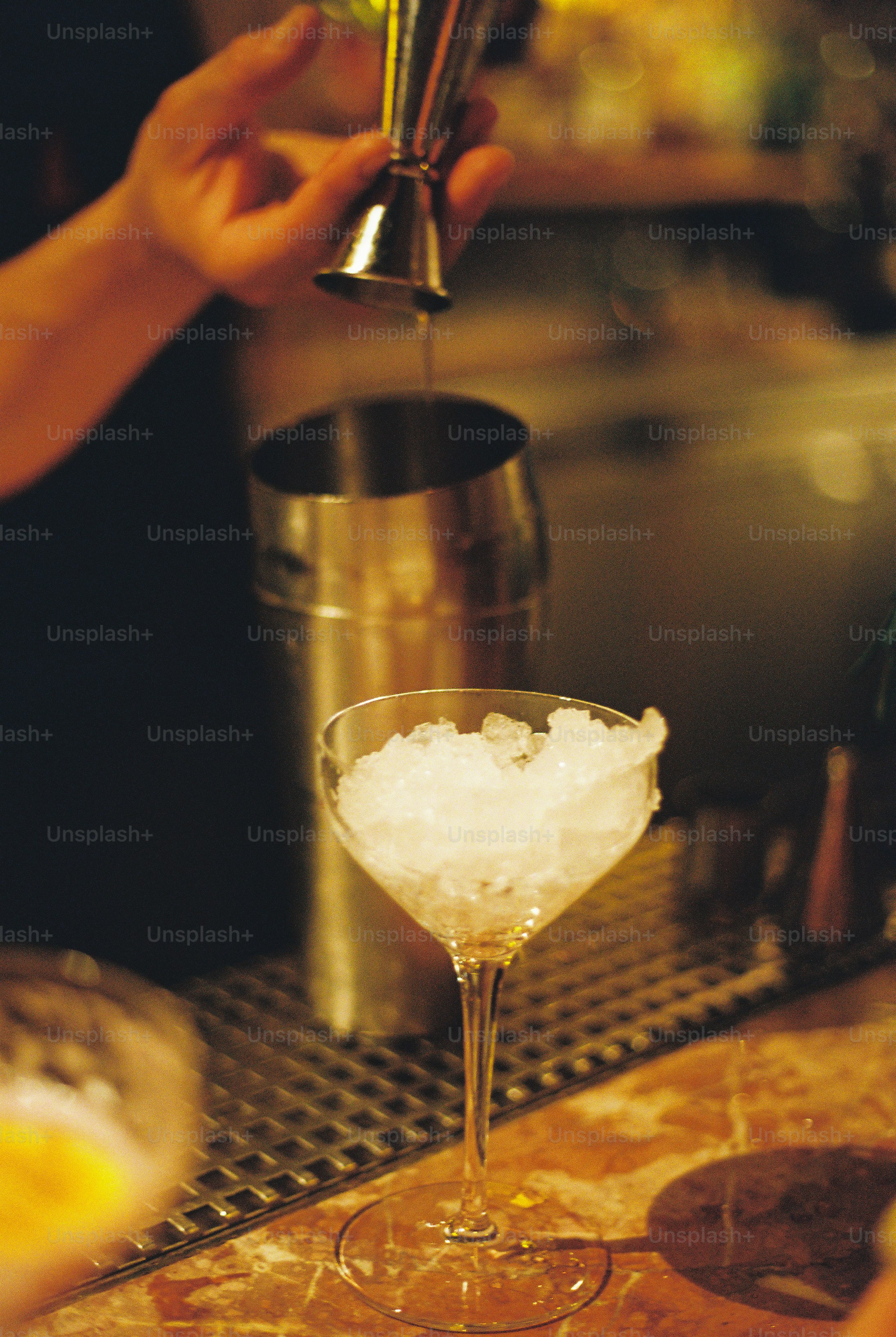 A hand pouring liquid from a jigger into a cocktail shaker near crushed ice