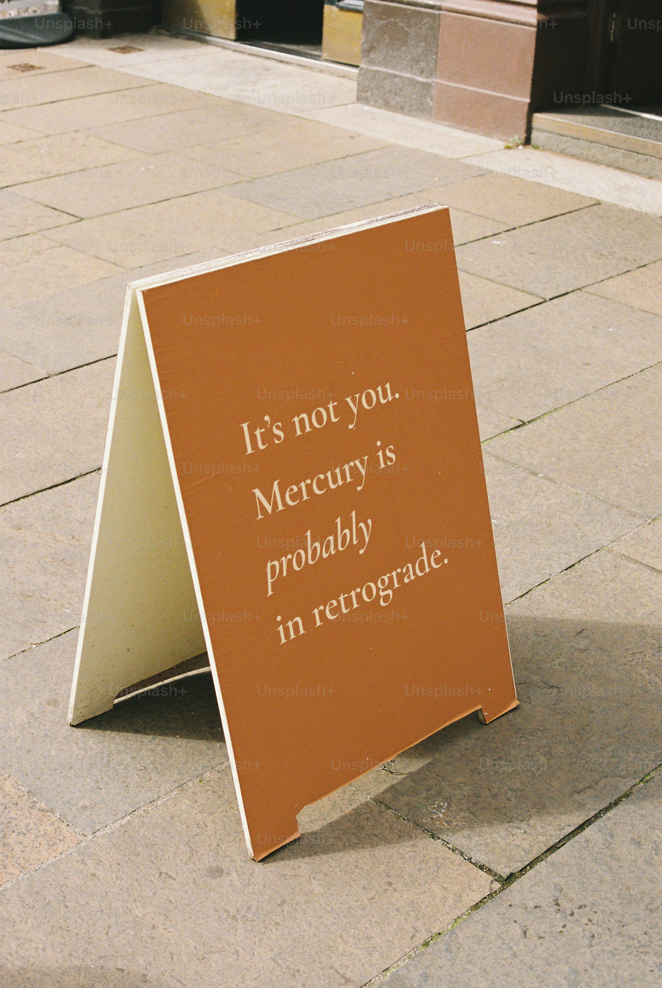 A brown sandwich board sign on a stone sidewalk with text about Mercury retrograde