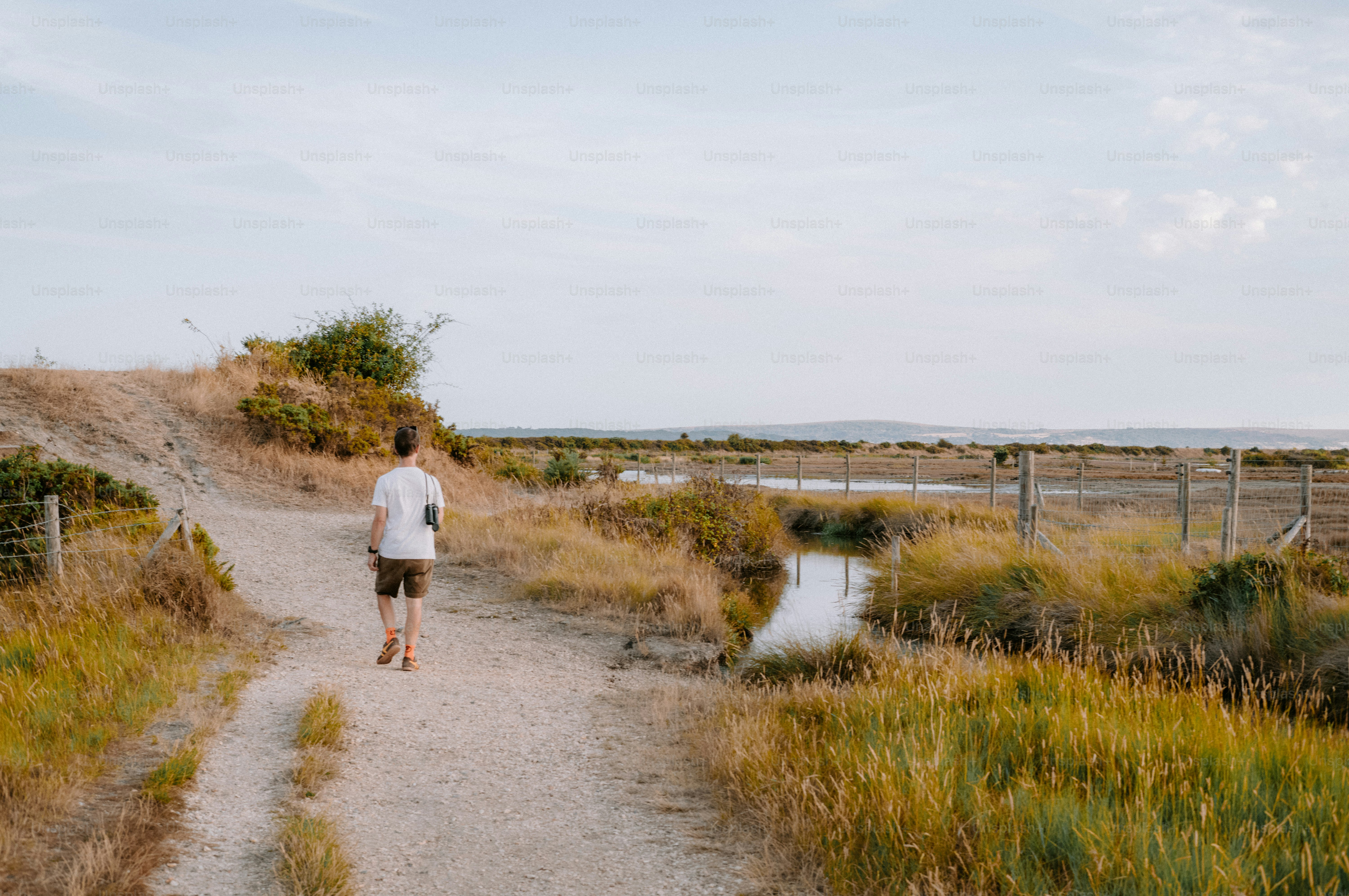 A person walking along a dirt path beside a narrow stream in a meadow