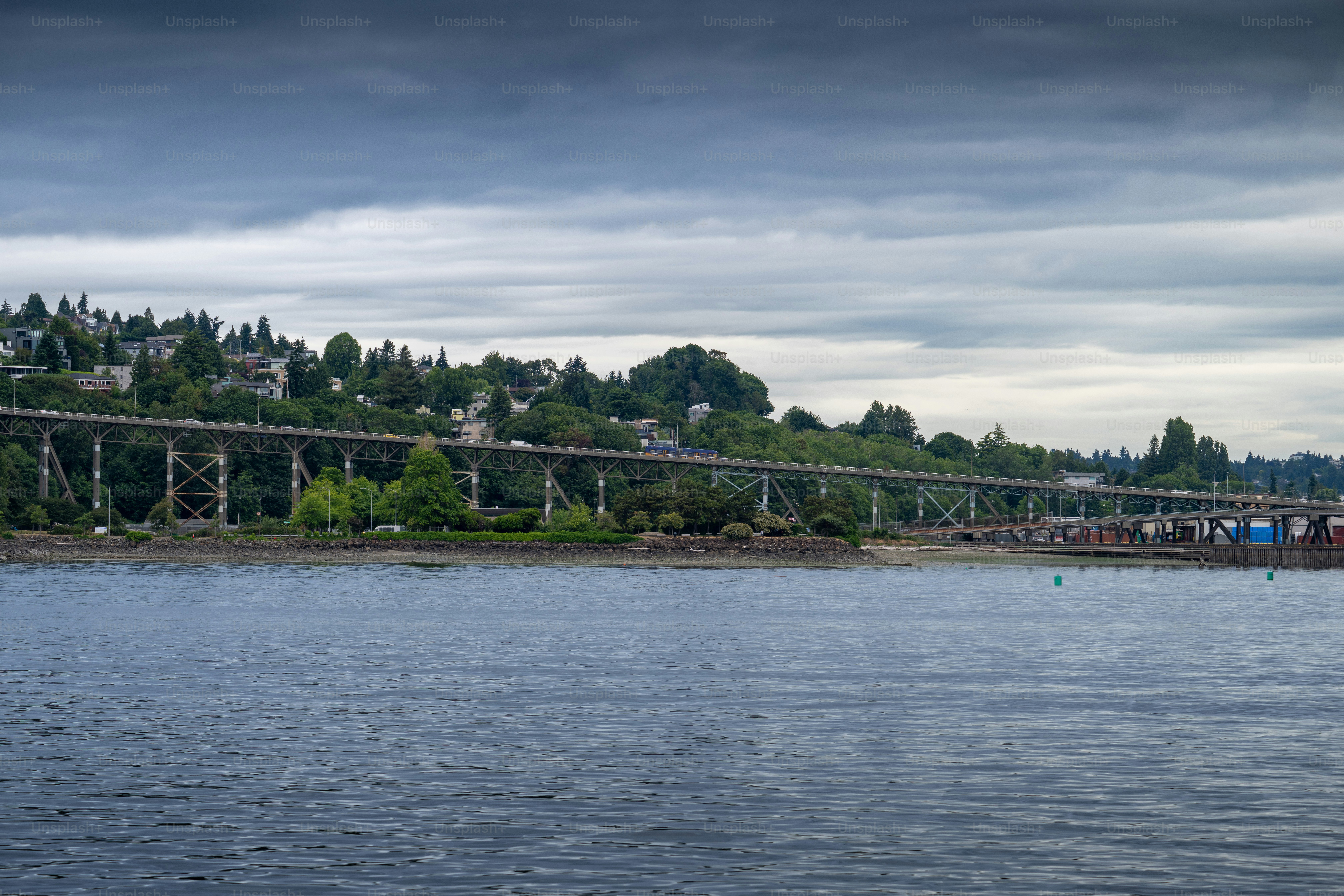 A wide view of the Aurora Bridge crossing Seattle’s Lake Union under dark, overcast skies. The bridge stretches above the water with lush green hills and houses in the background.