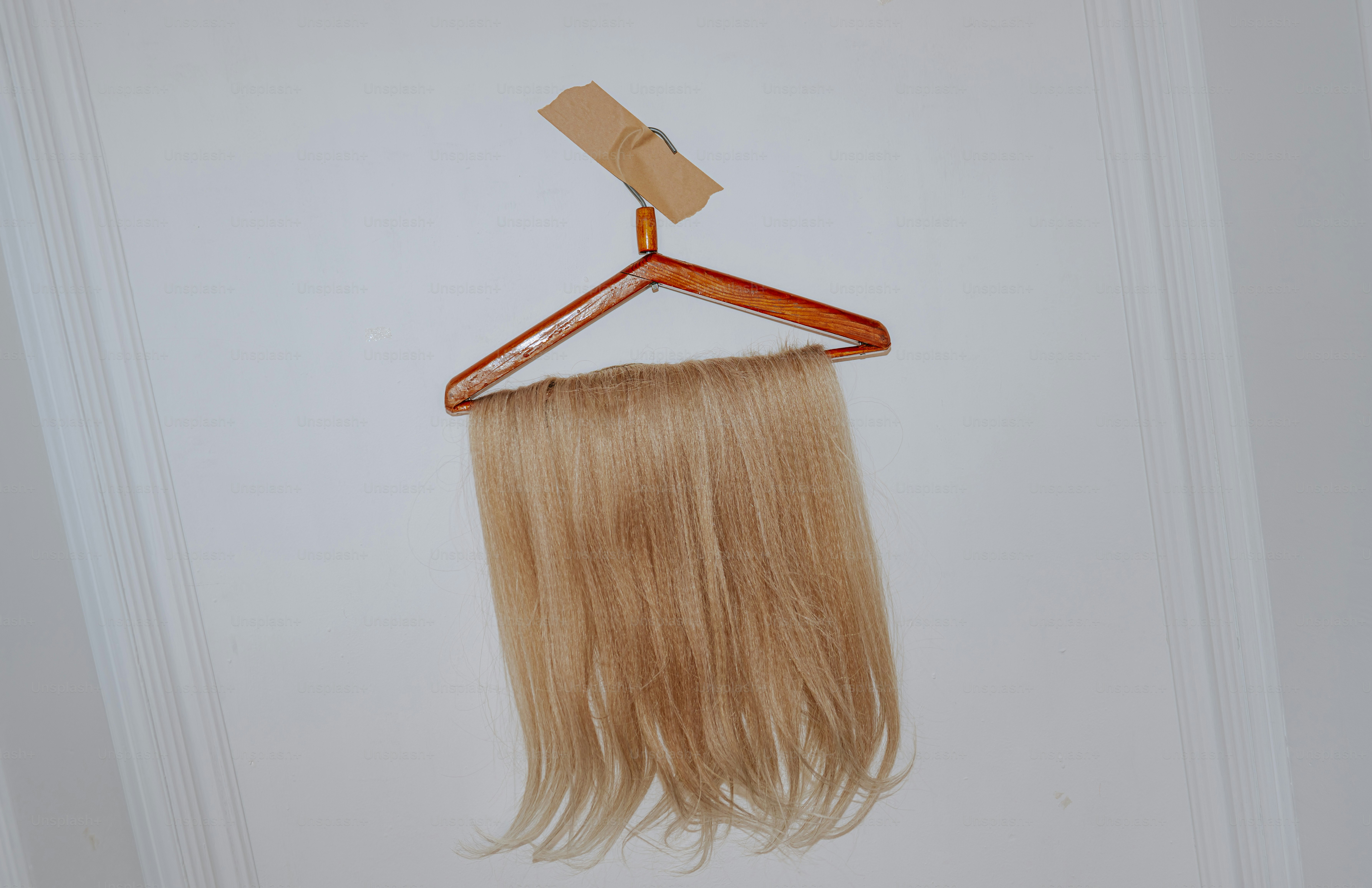 A blonde hair extension hanging from a wooden clothes hanger taped to a wall