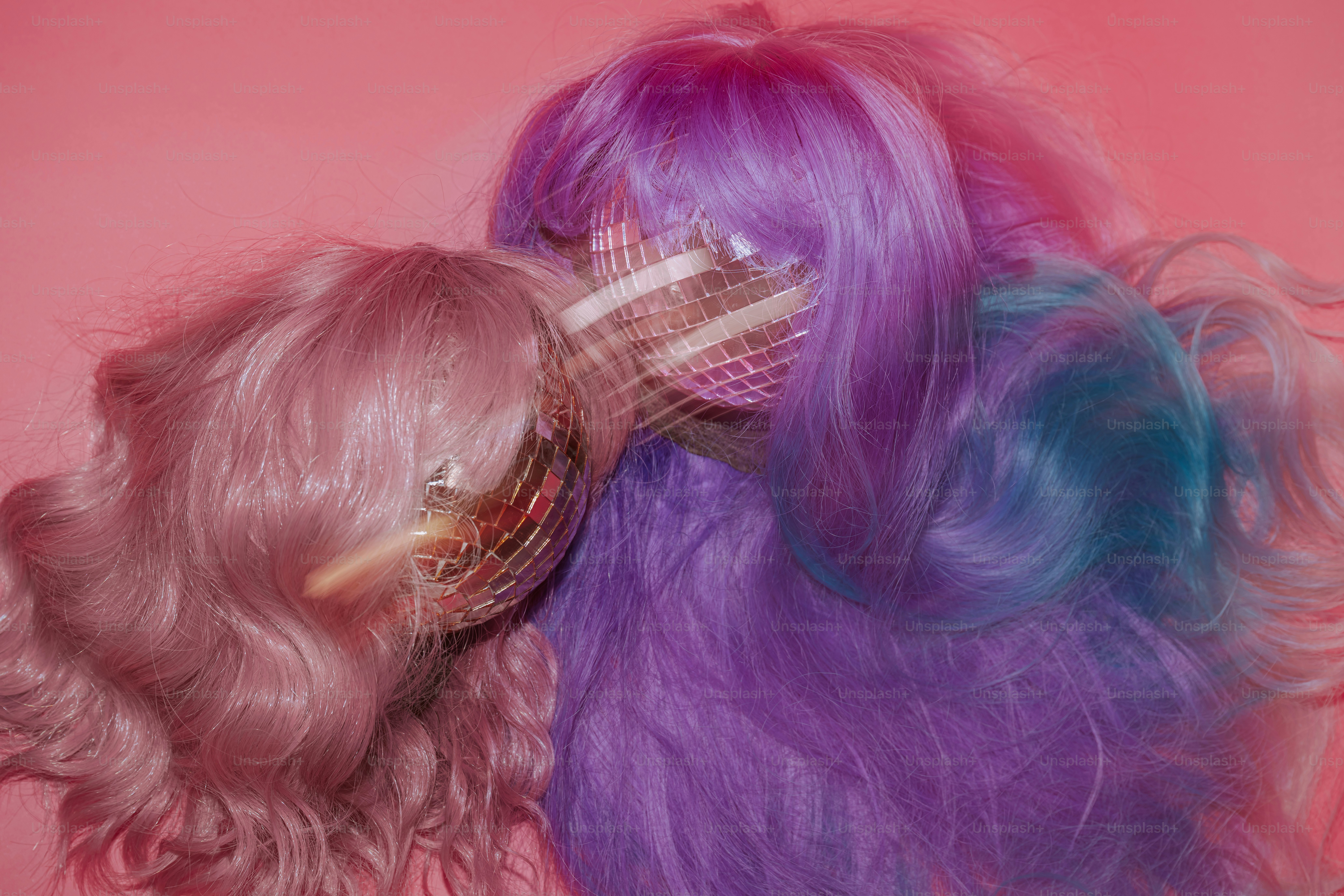 Two disco balls partially covered by pink and purple wigs against a pink background