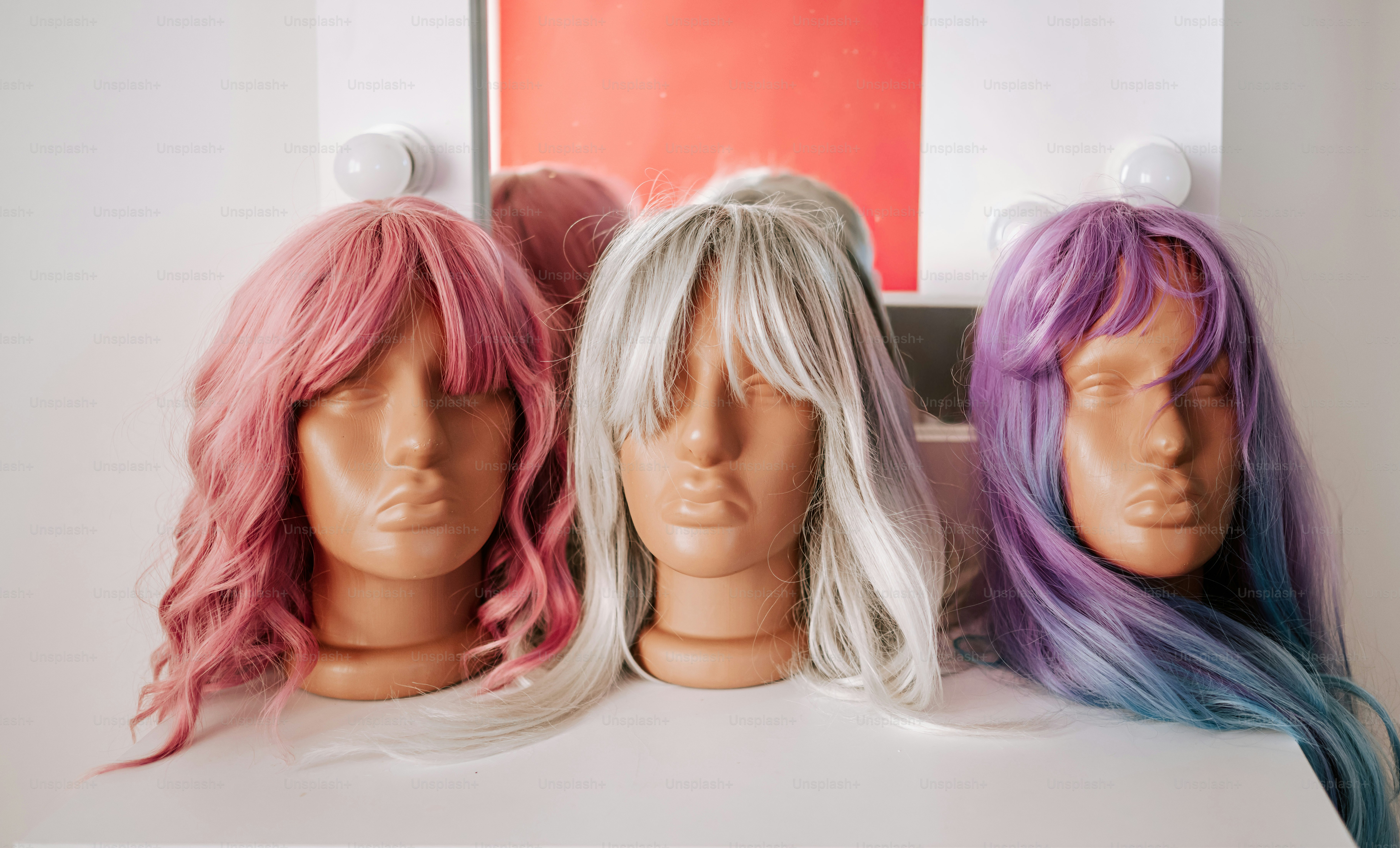 Three mannequin heads wearing pink, silver, and purple wigs on a white table