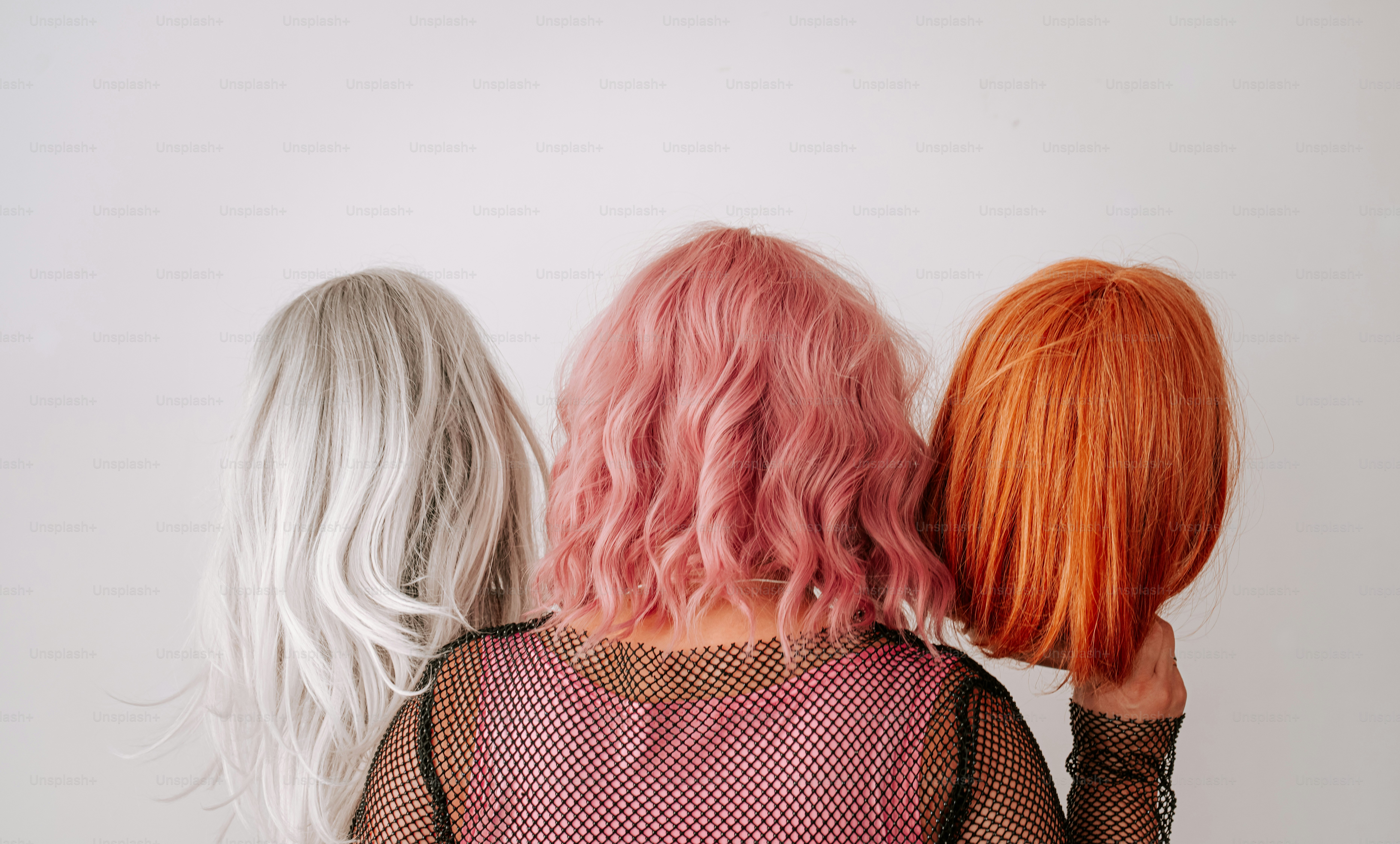 A person in a fishnet top holding three colorful wigs against a white background