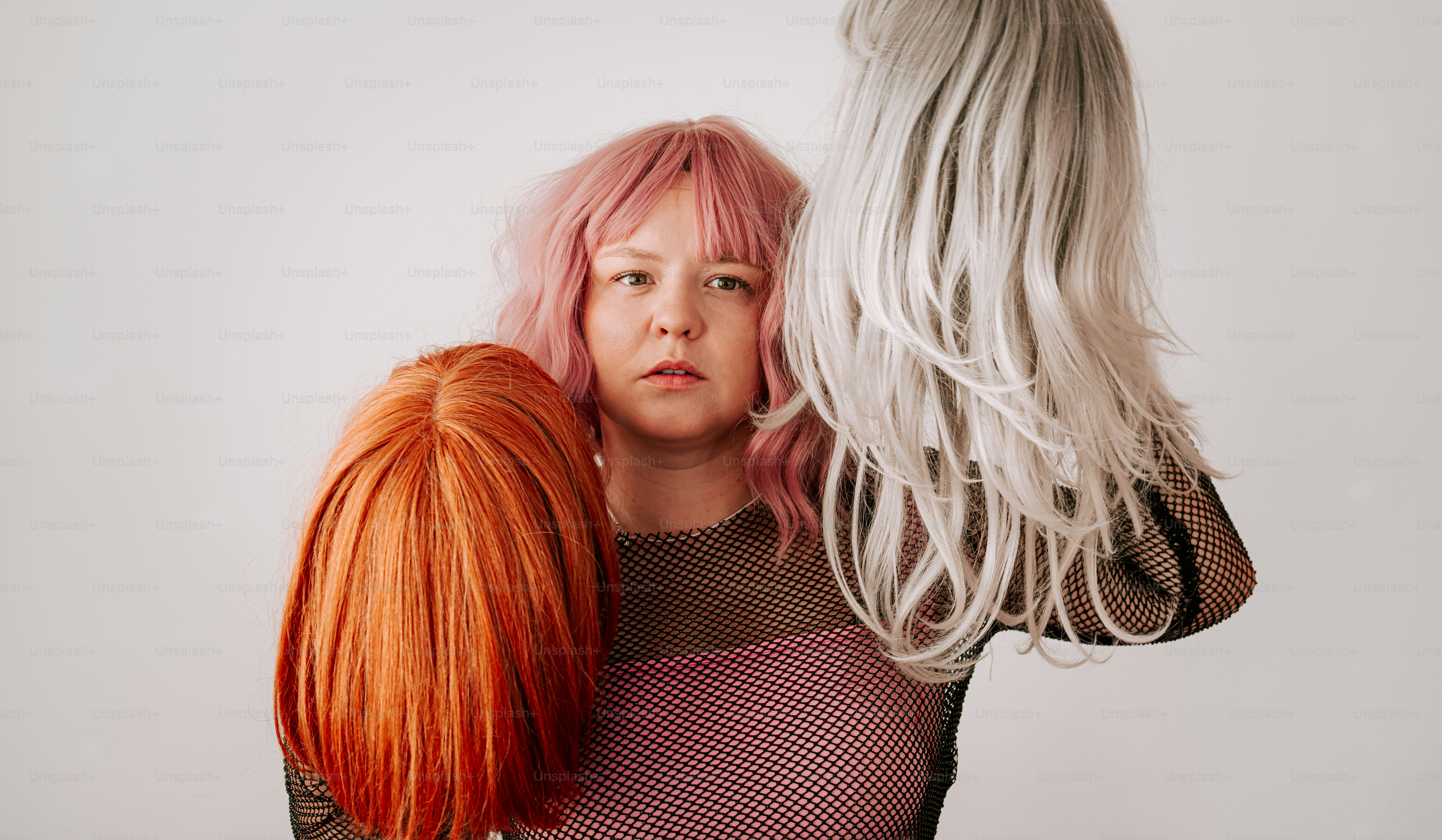 A person with pink hair holding an orange wig and a silver wig