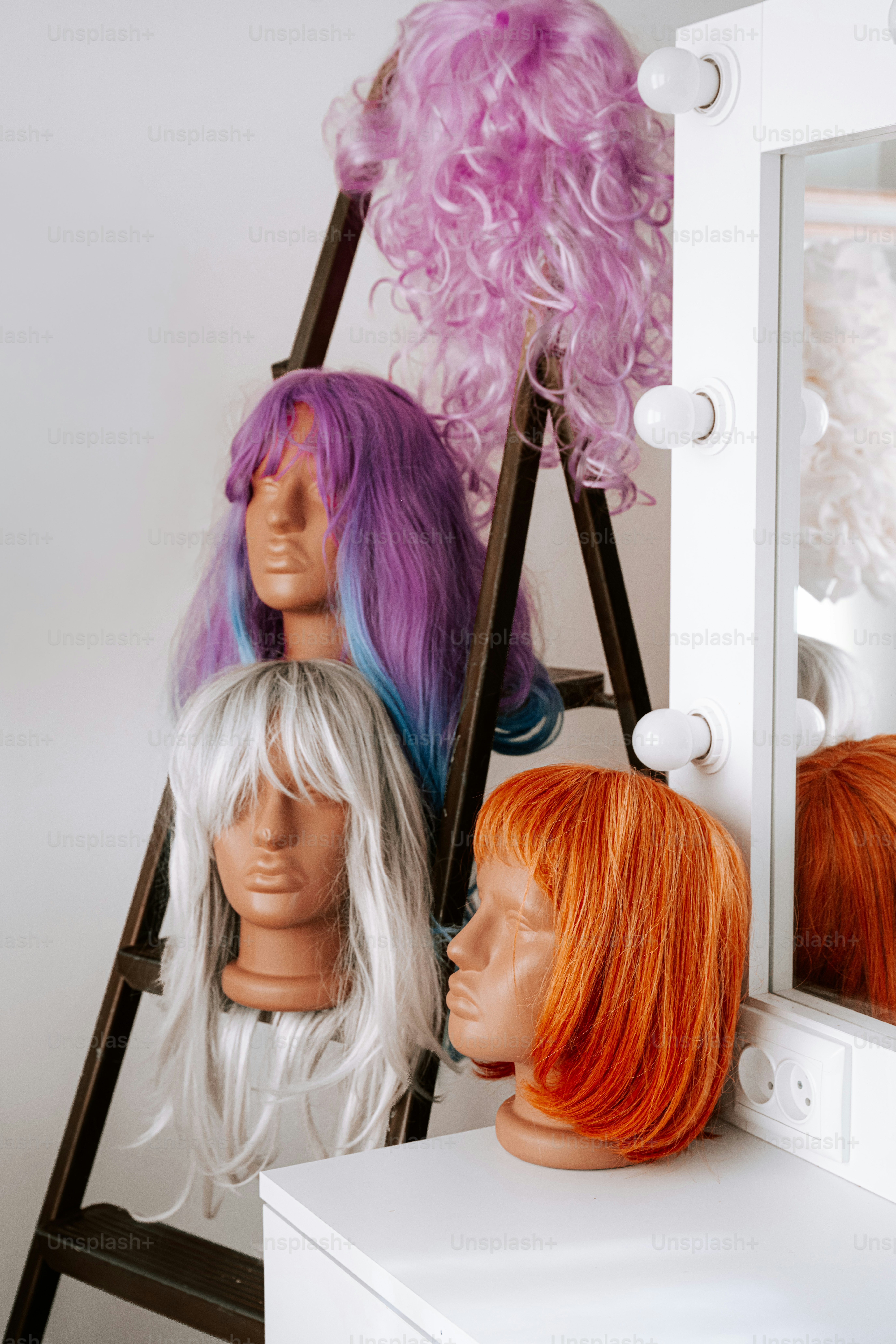 Three mannequin heads wearing colorful wigs on a ladder beside a vanity mirror