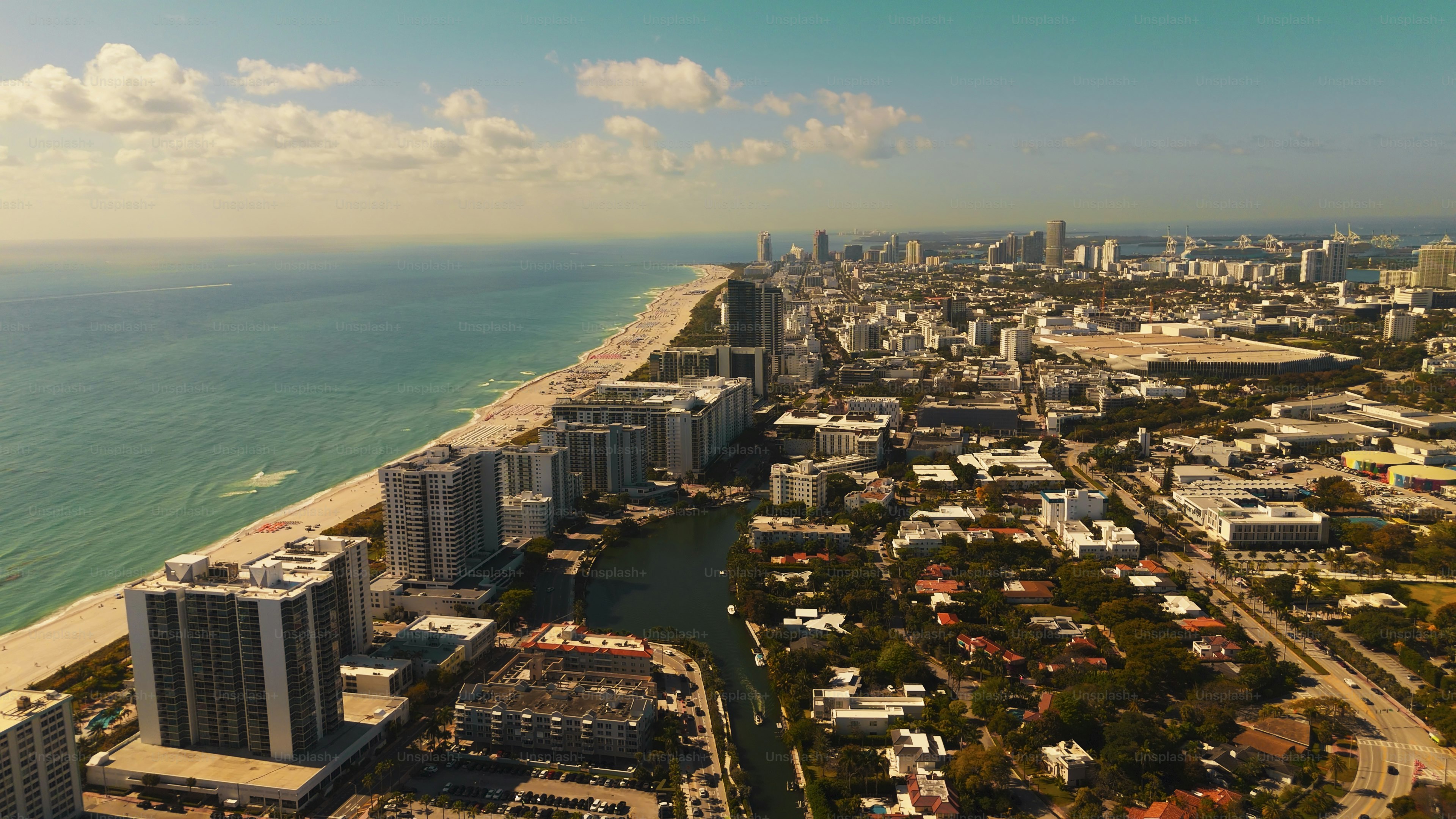Aerial view of Miami's beachfront extending northward along turquoise waters, with high-rise residential towers lining the sandy shore and downtown Miami's illuminated skyline visible in the distance under warm afternoon light.