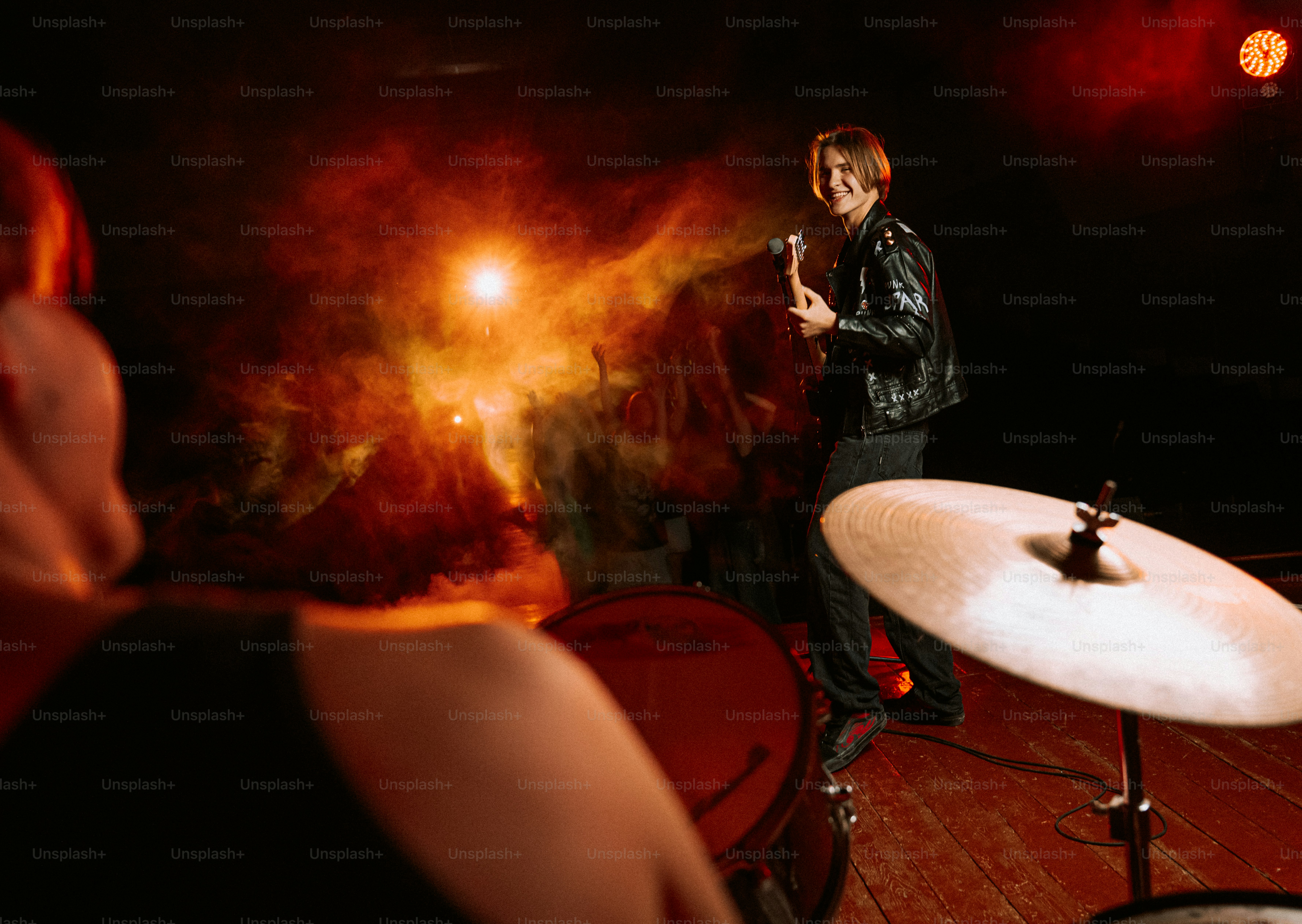 A guitarist in a leather jacket performing on a stage with orange smoke