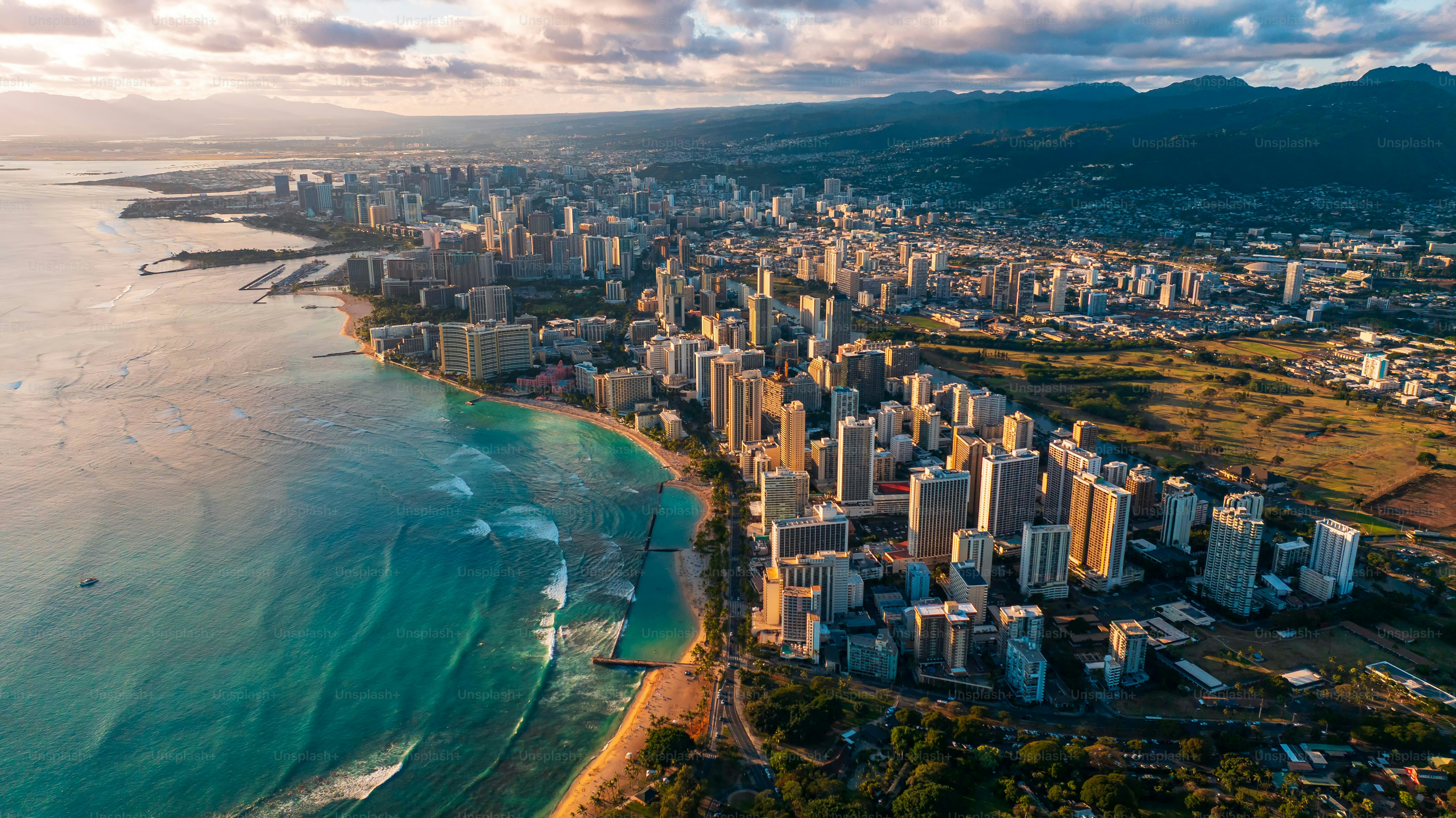 Urban landscape of Waikiki resort is lit with setting sun. Ocean waves roll to the sandy beaches. Dark mountains at backdrop are under grey clouds. Aerial view.