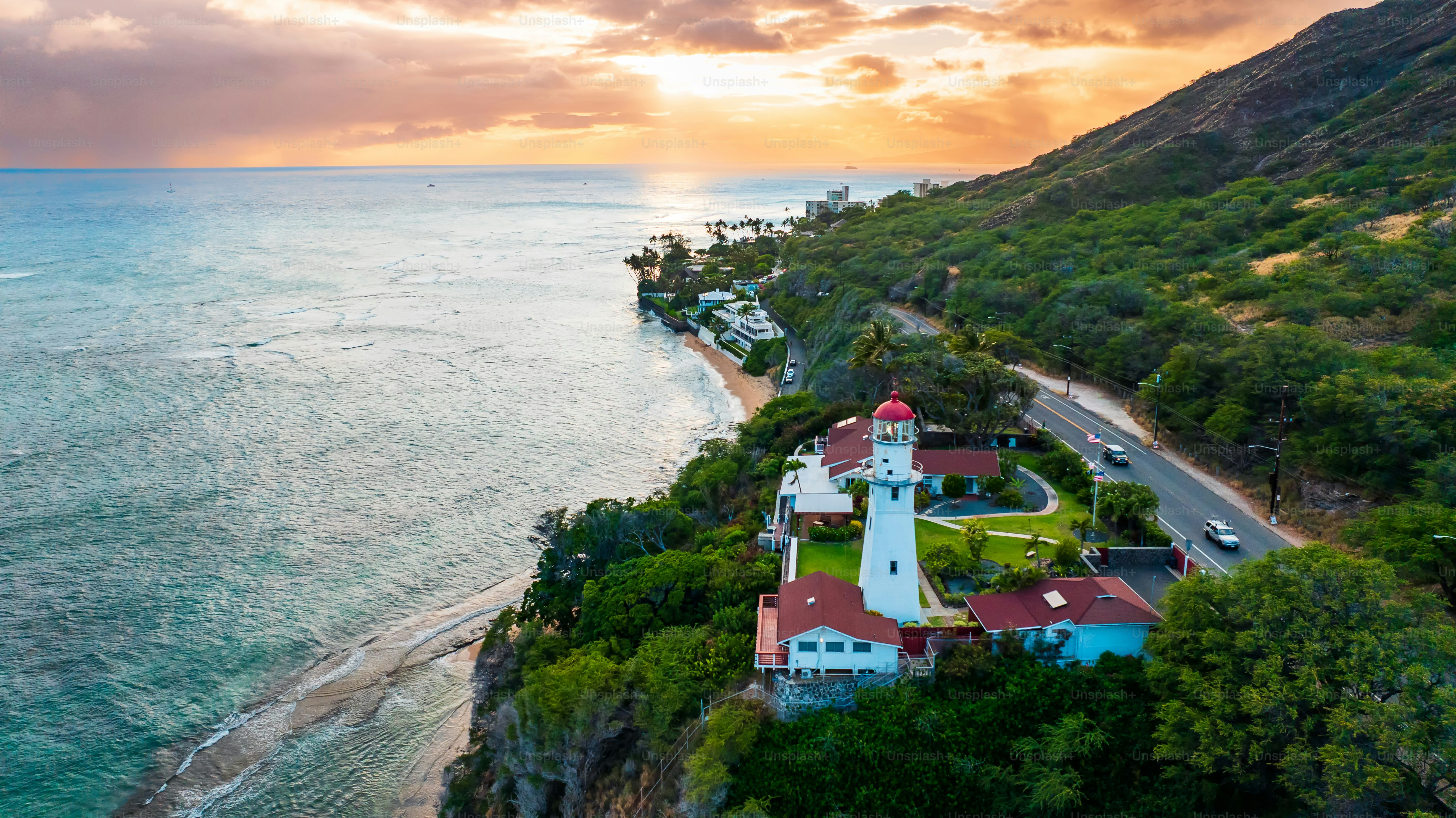 White lighthouse on the picturesque coast at the mountain foot. Aerial view on the Pacific Ocean waterscape at sunset.