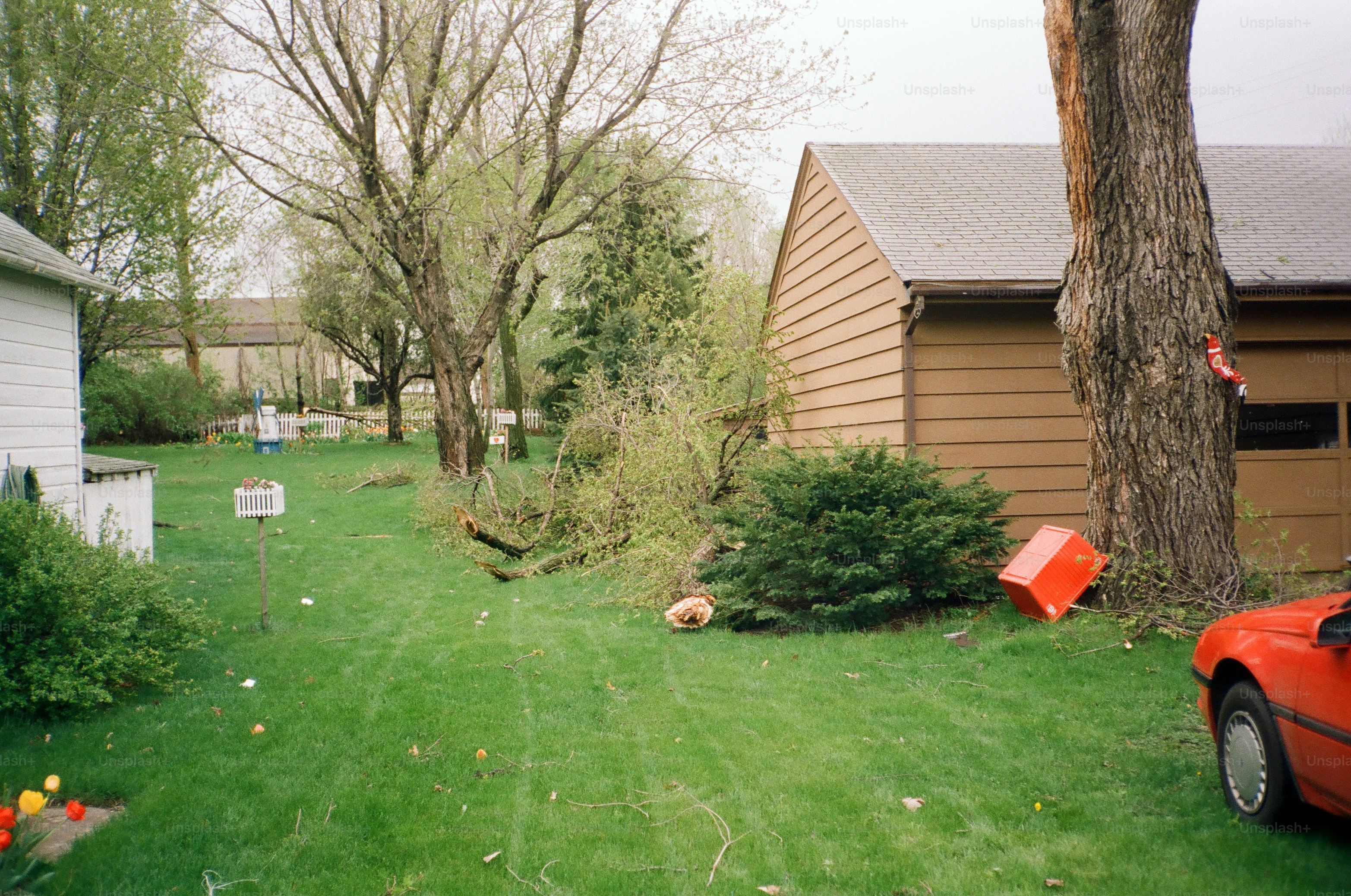A suburban backyard showing aftermath of a windstorm with fallen tree limbs, scattered debris, and overturned objects near a garage and green lawn.