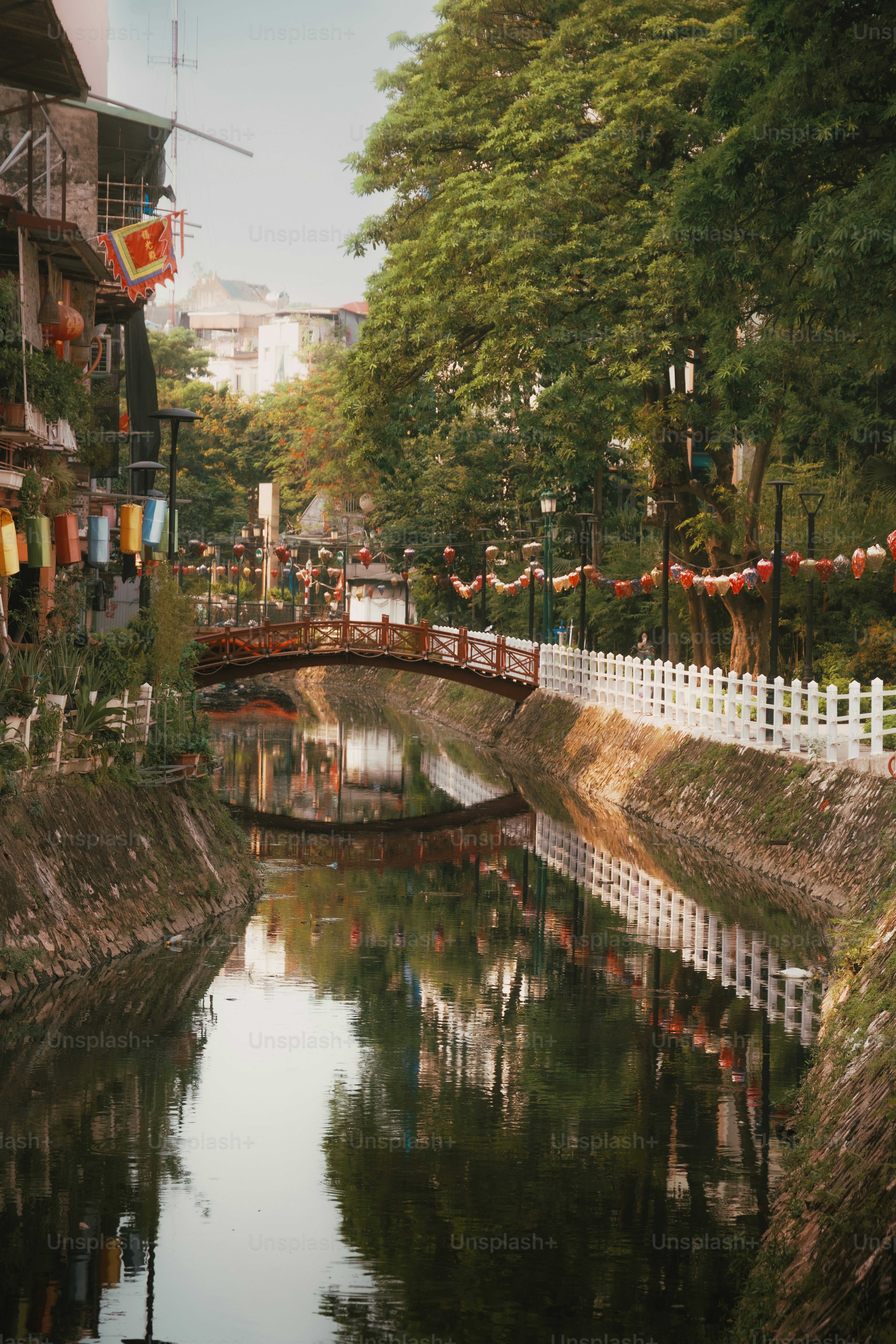 Uma ponte de madeira arqueada sobre um canal estreito ladeado por árvores e lanternas