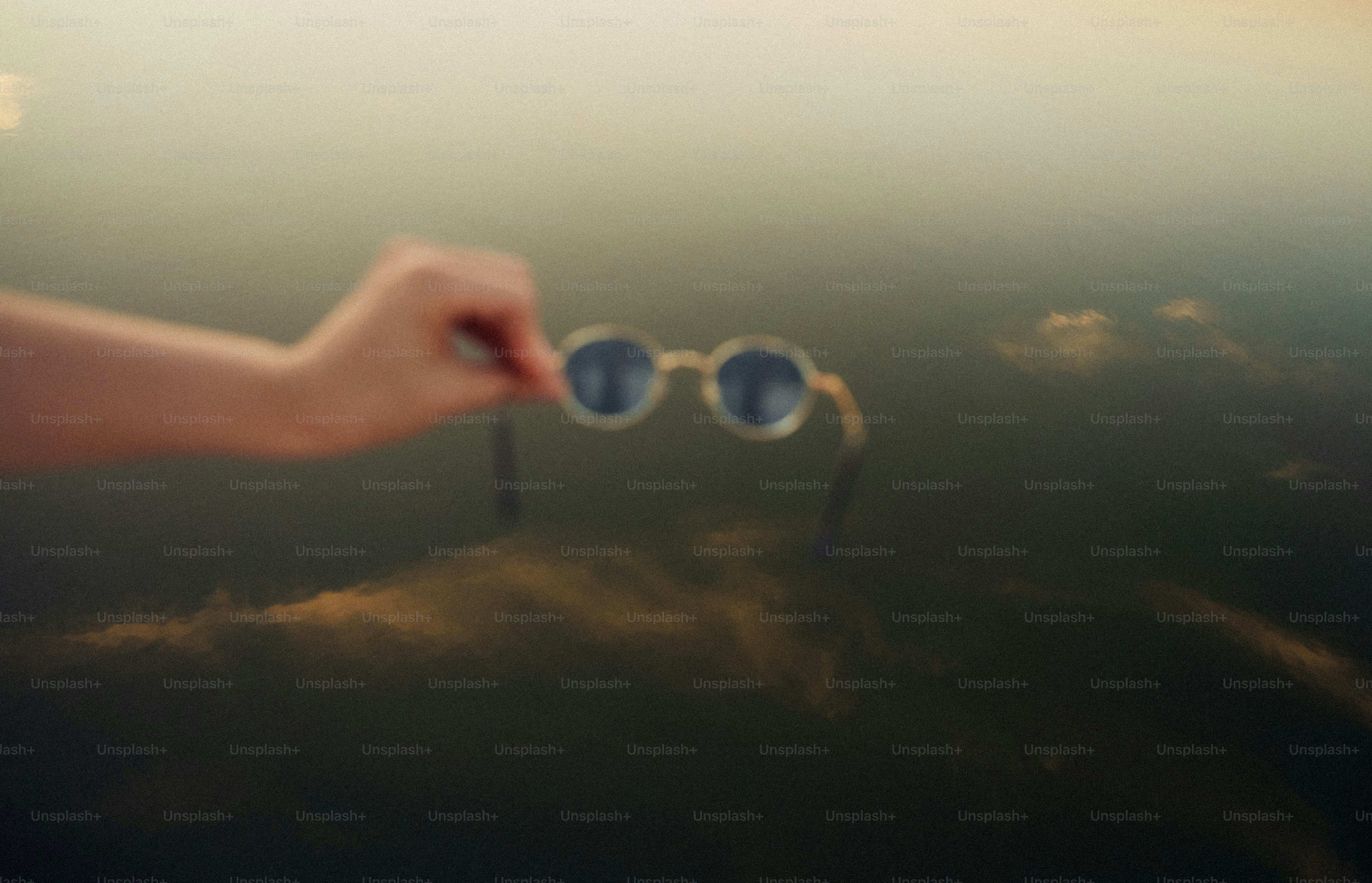 A hand holding round sunglasses against a blurred reflection of clouds on water