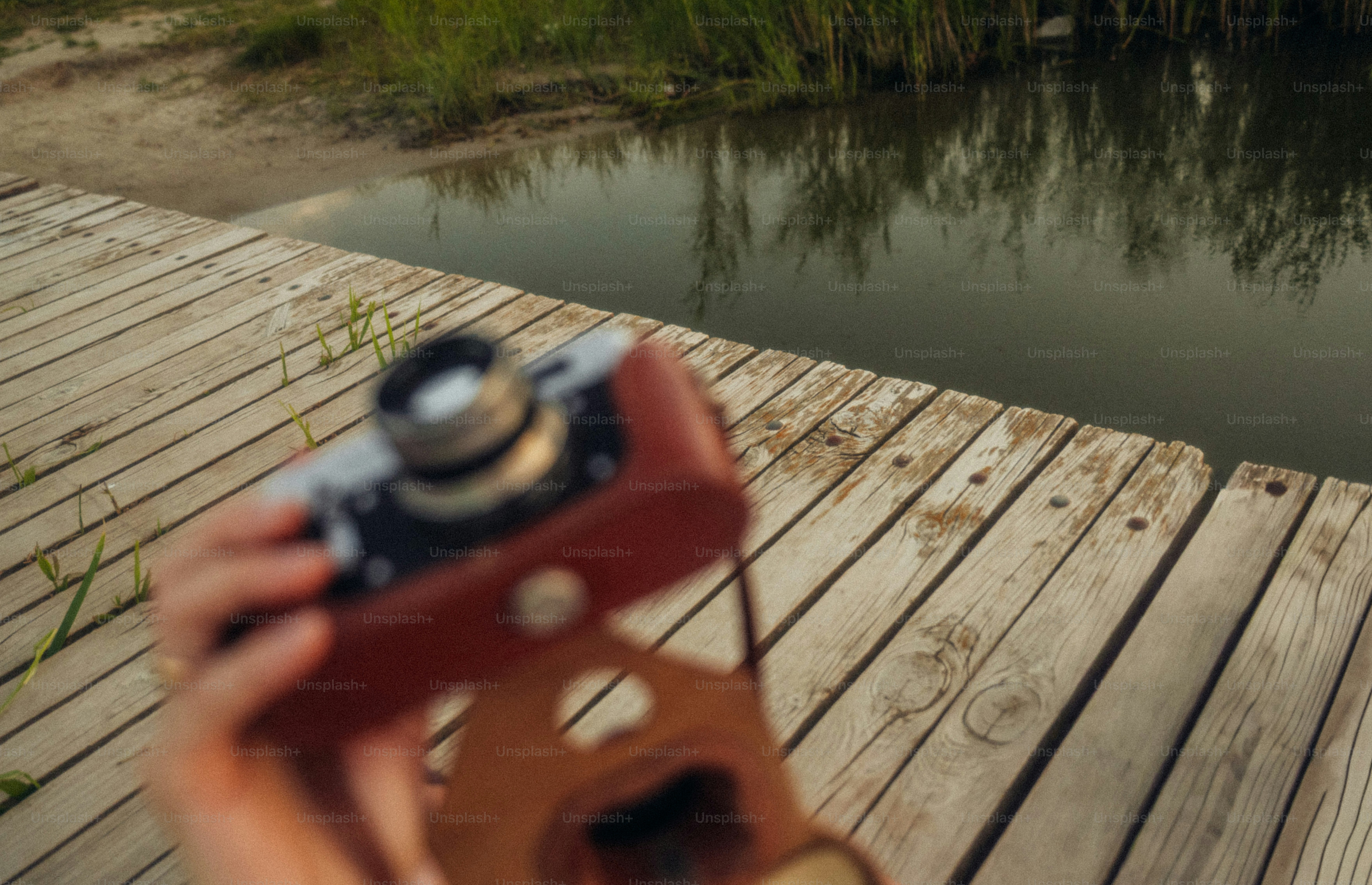 A person holding a vintage camera over a wooden pier by a lake