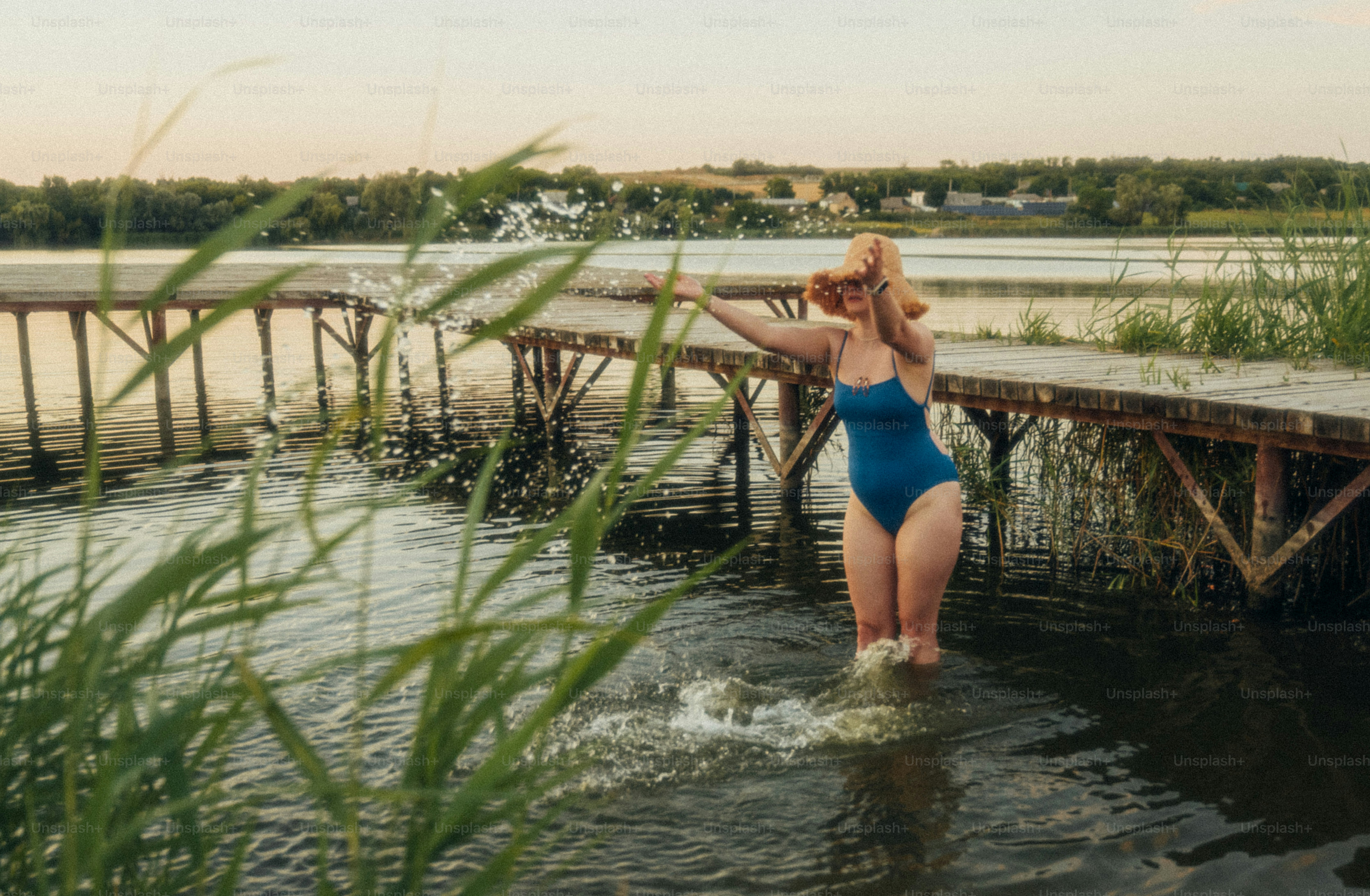 A woman in a blue swimsuit and straw hat splashing water near a wooden pier