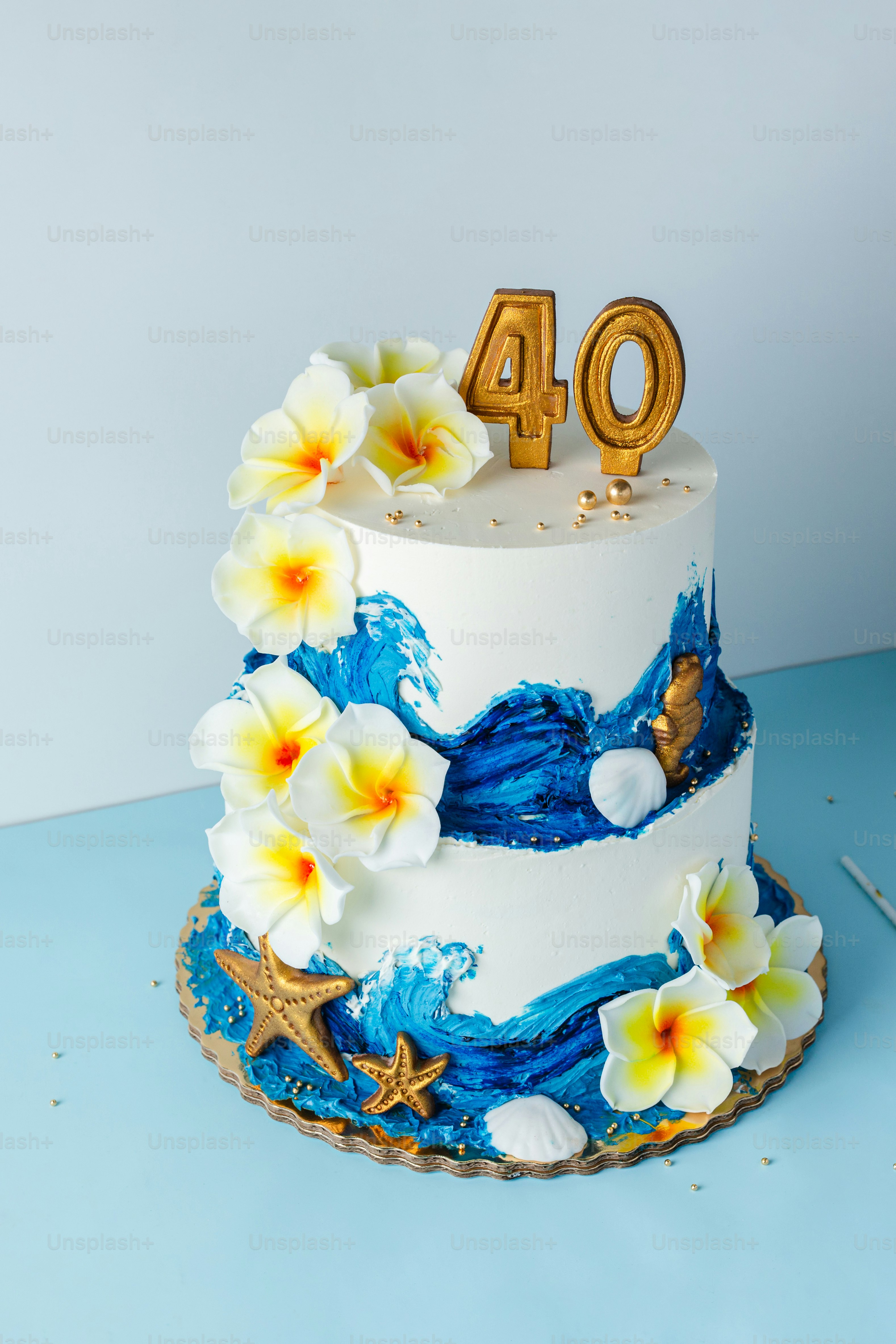 Una torta a due piani decorata con onde blu, fiori di plumeria bianchi e stelle marine dorate