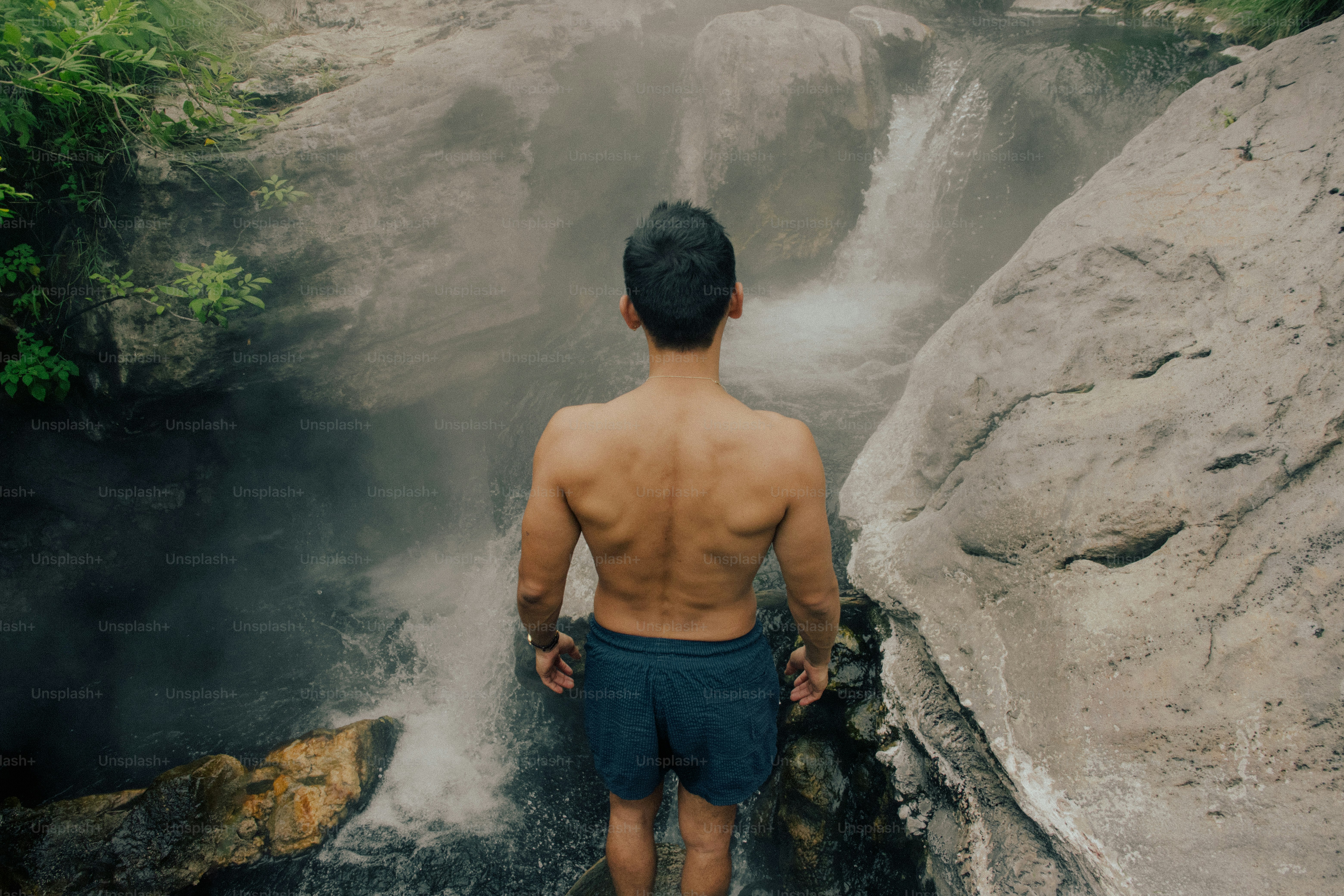 Un uomo a torso nudo in pantaloncini scuri che si trova di fronte a una cascata nebbiosa tra scogliere rocciose