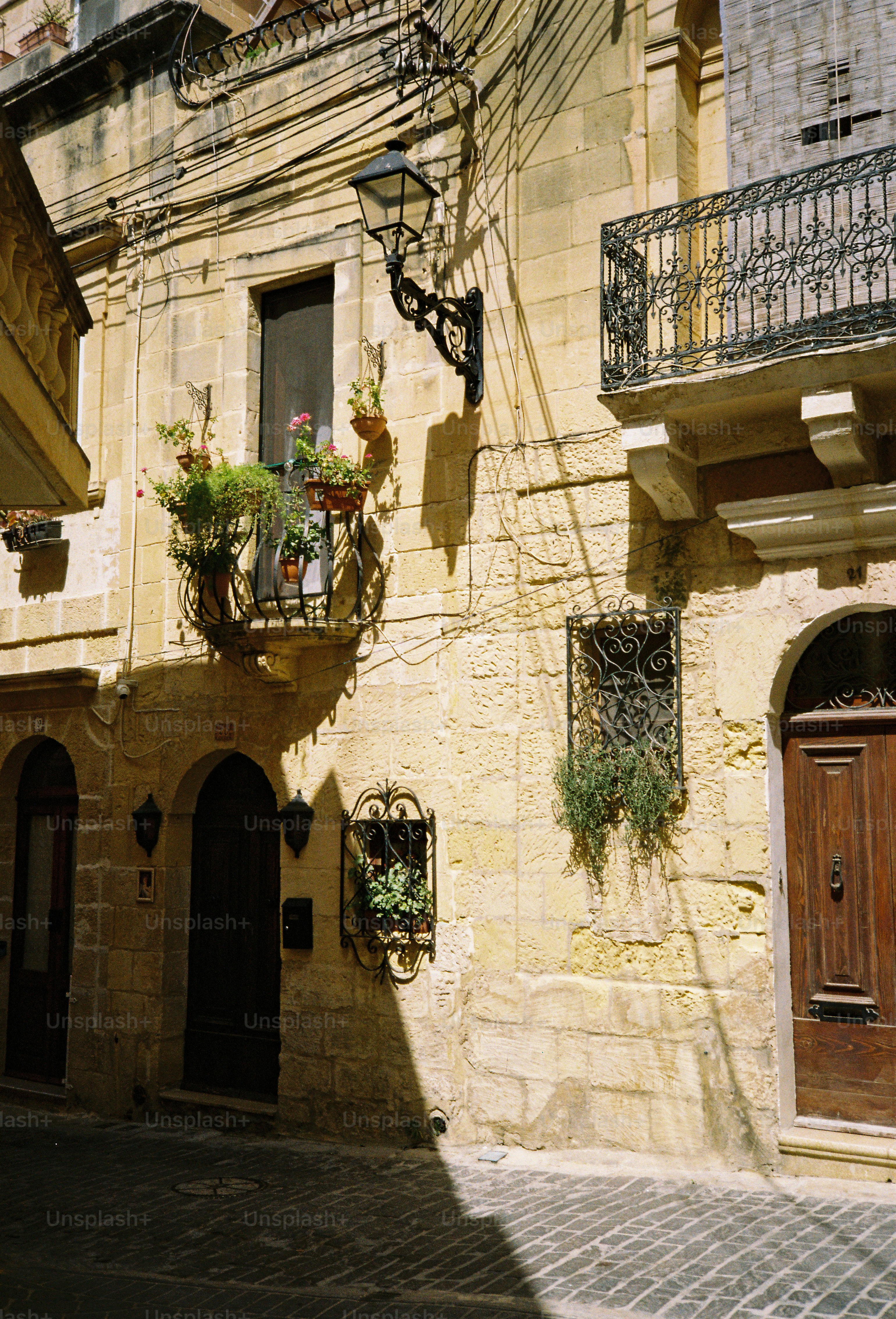A sunlit limestone building with a wrought iron balcony and potted plants