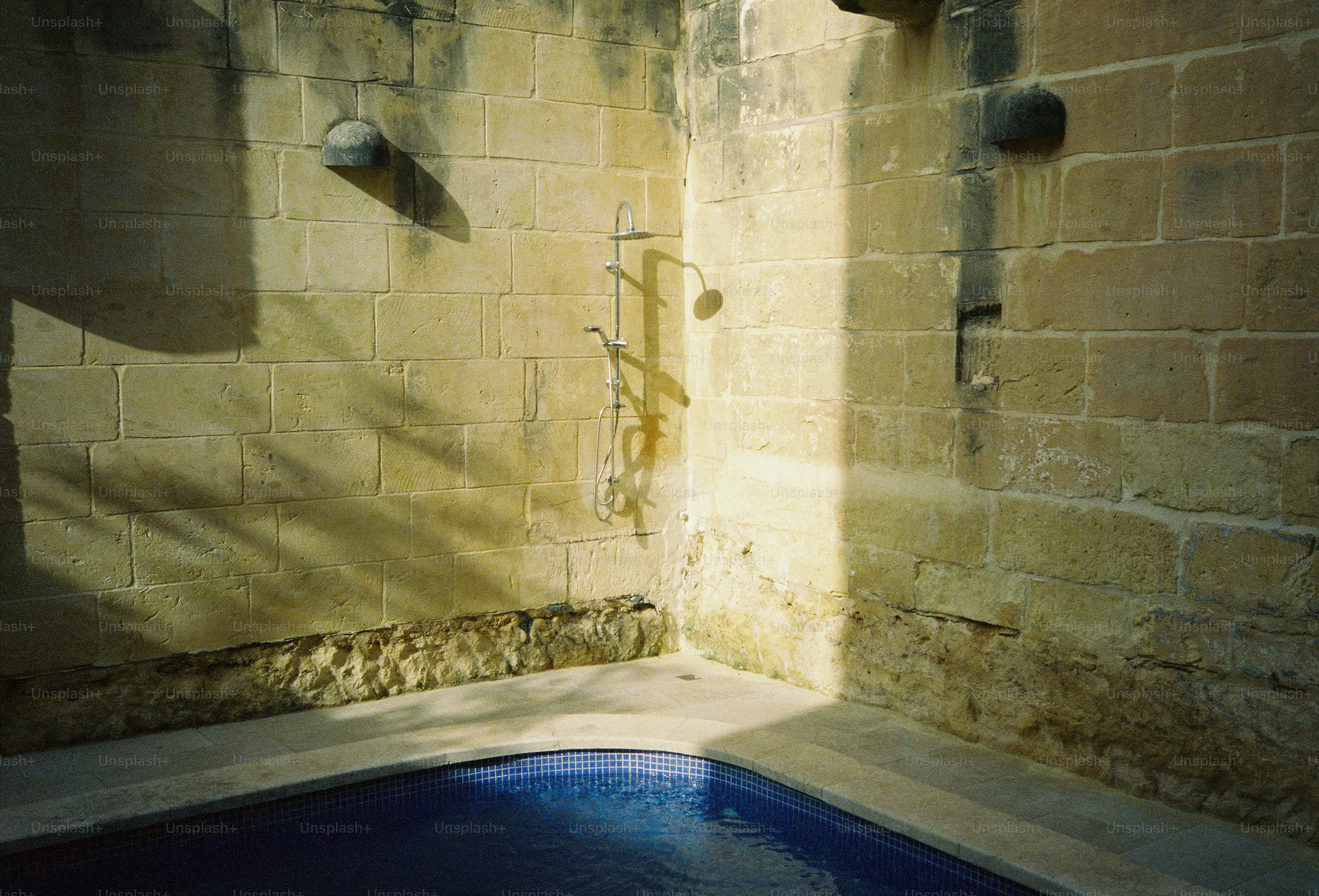 A chrome shower fixture mounted on a weathered stone wall above a blue pool