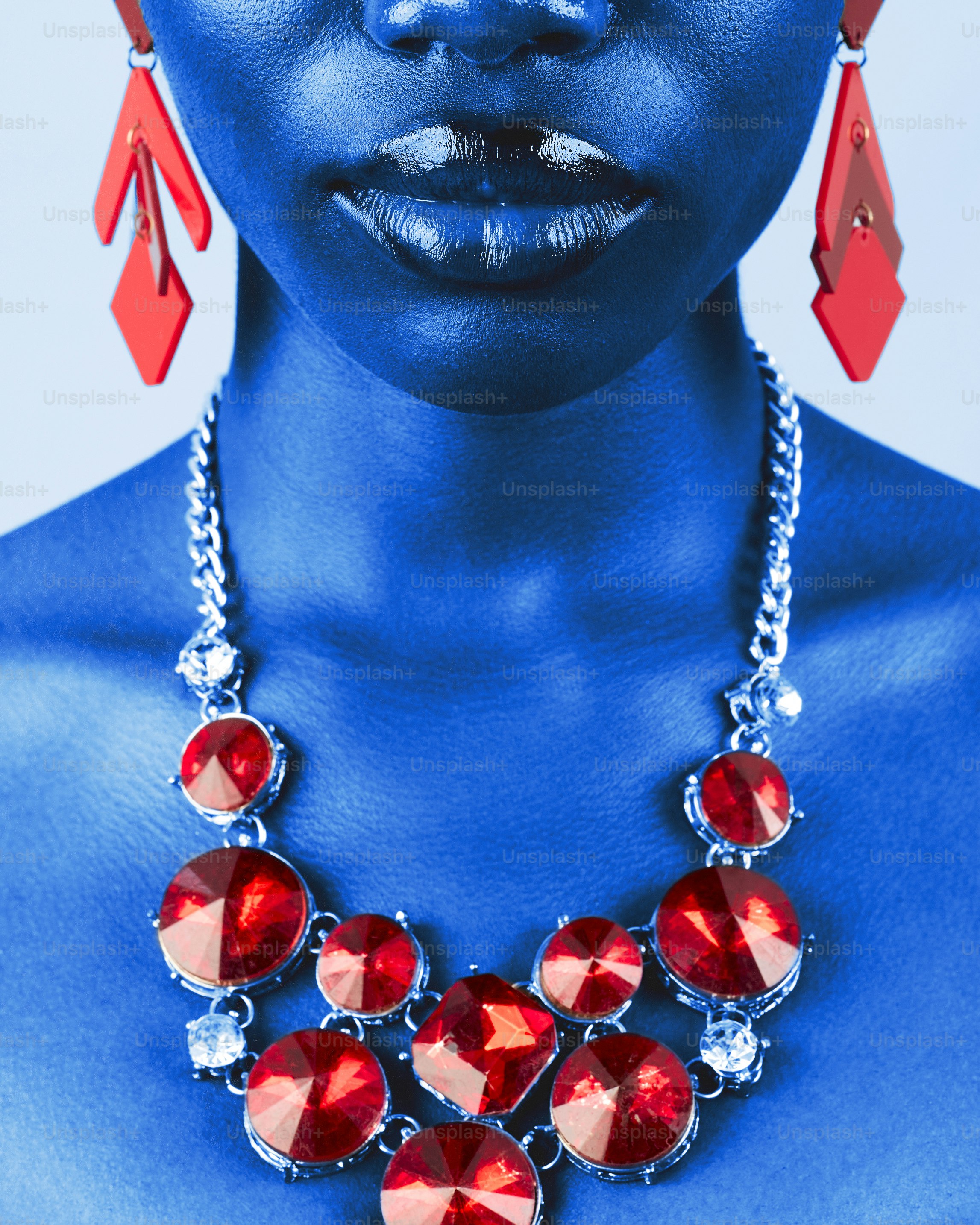 Uma pessoa de pele azul usando brincos geométricos vermelhos e um colar de cristal vermelho