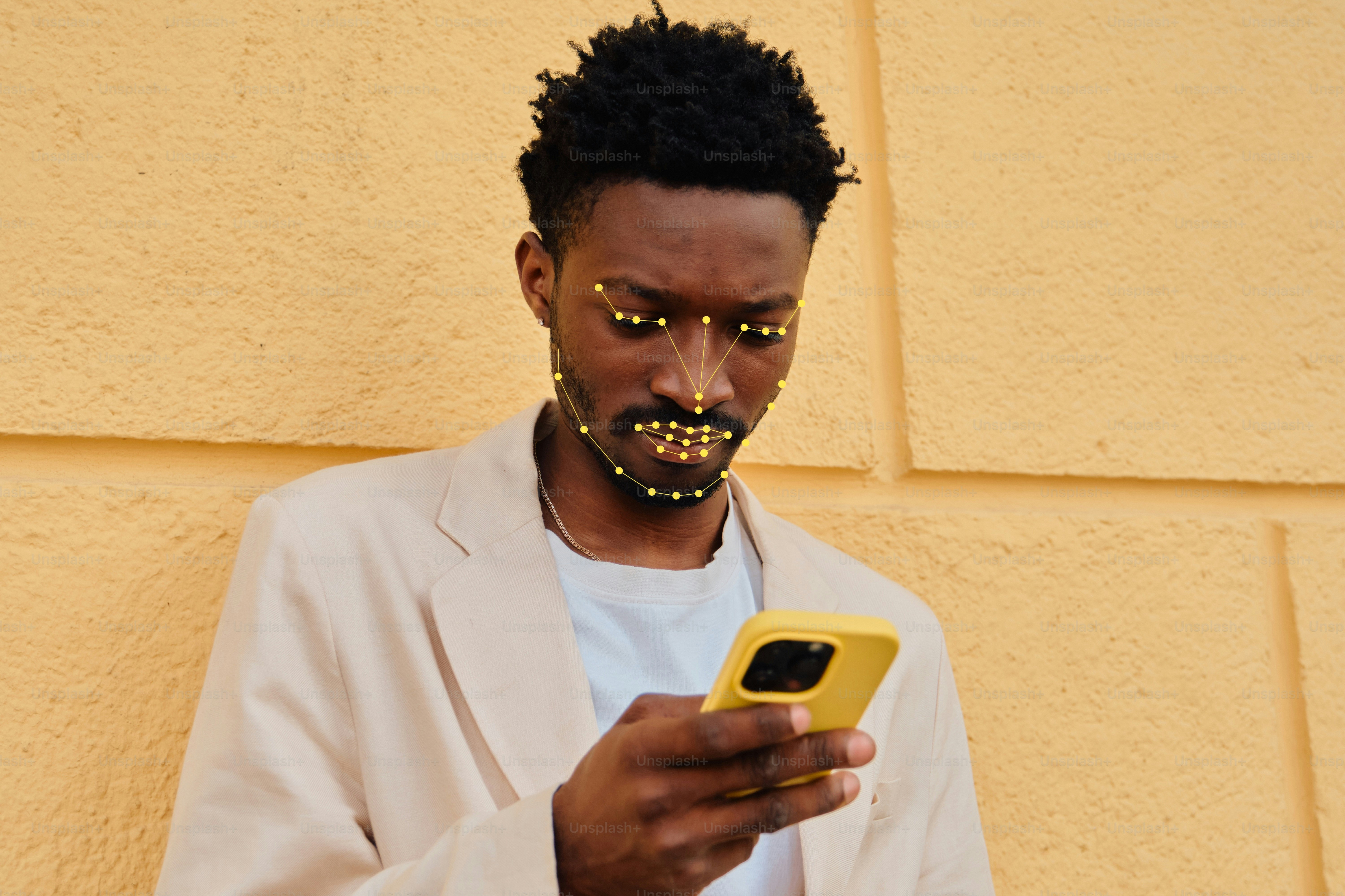 A man with facial mapping dots looking at a yellow smartphone against a yellow wall