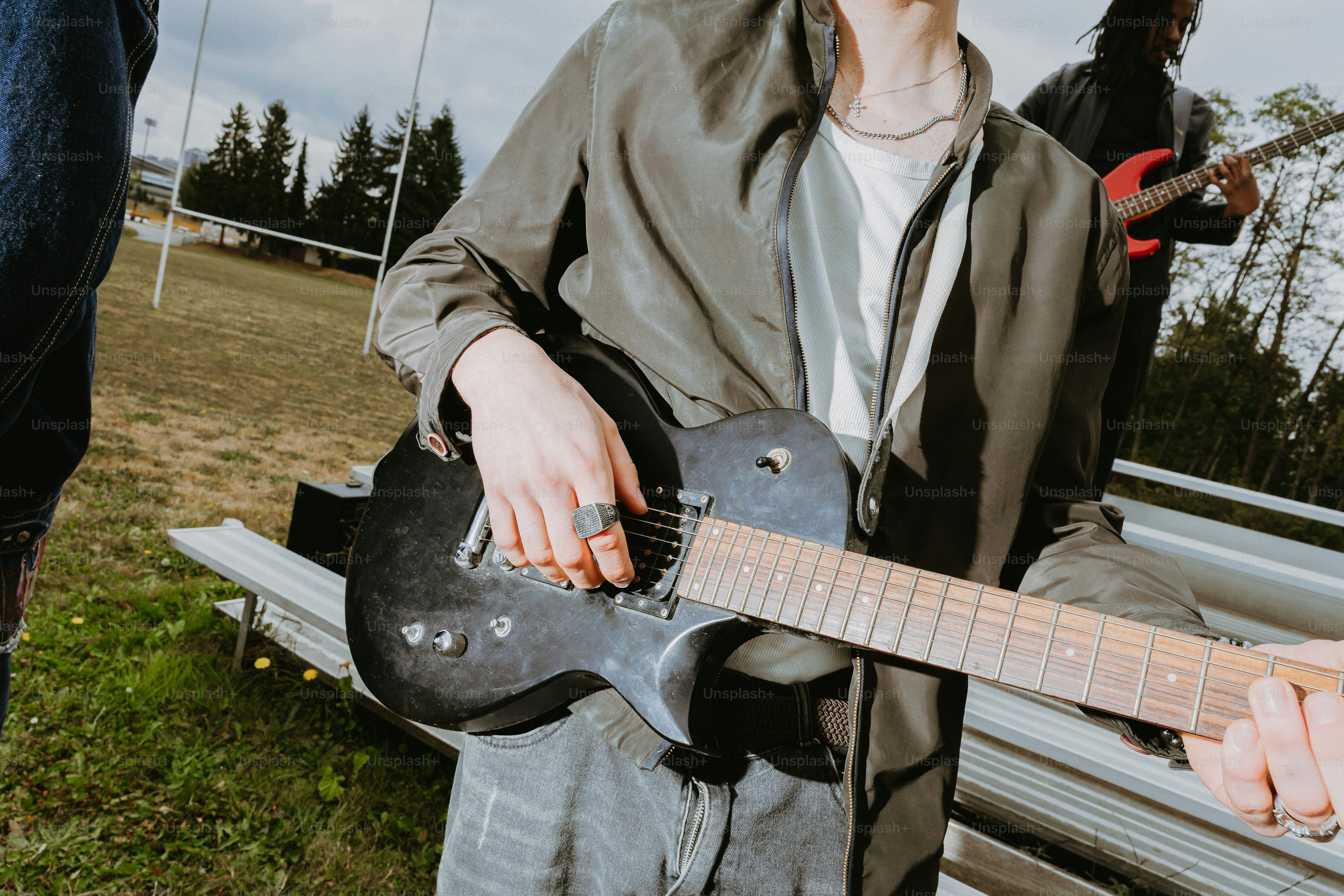 Una persona che suona una chitarra elettrica nera all'aperto con un altro musicista sullo sfondo