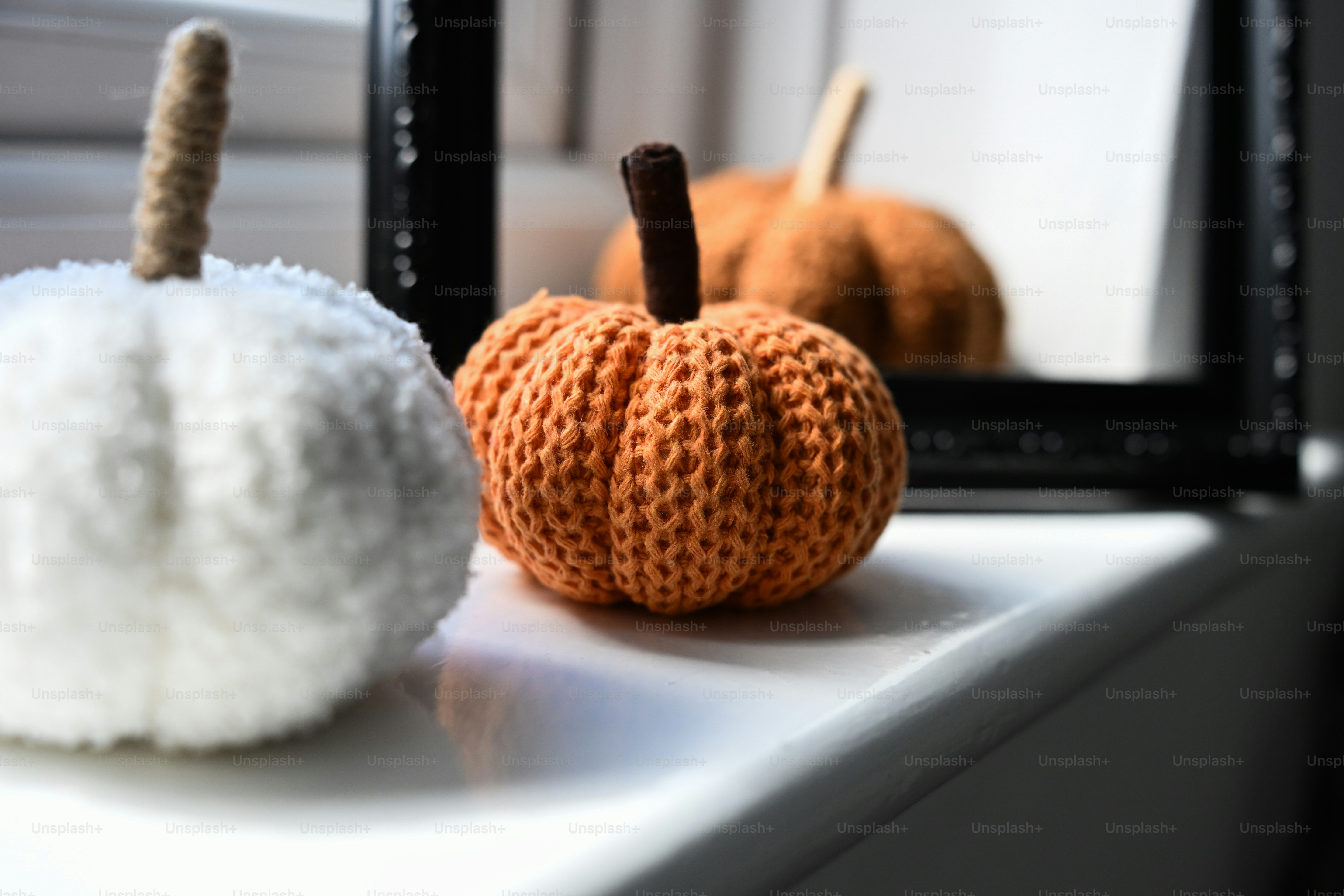 A white and an orange knitted pumpkin on a white windowsill