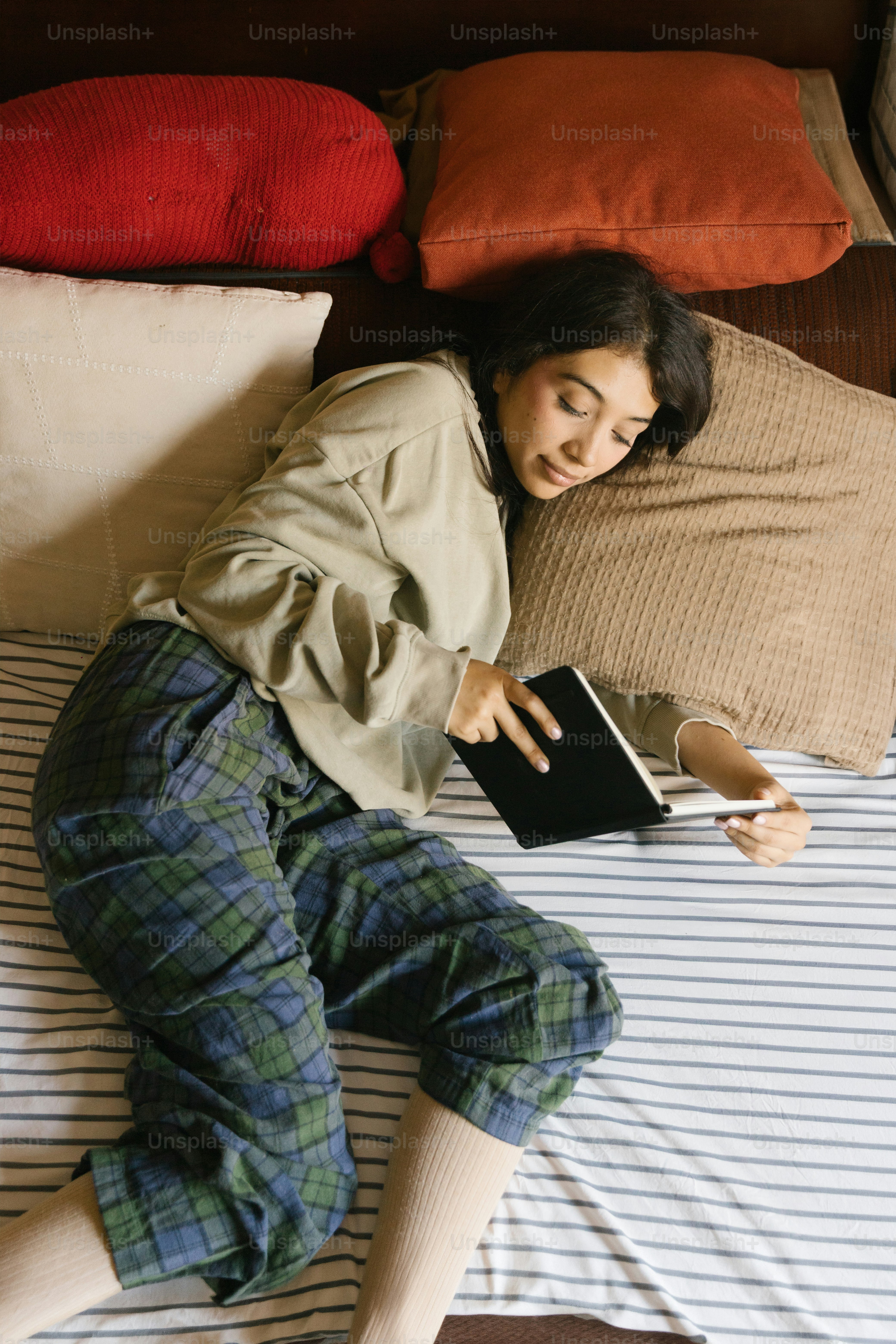 A woman in a beige sweatshirt and plaid pants reading a book on a bed