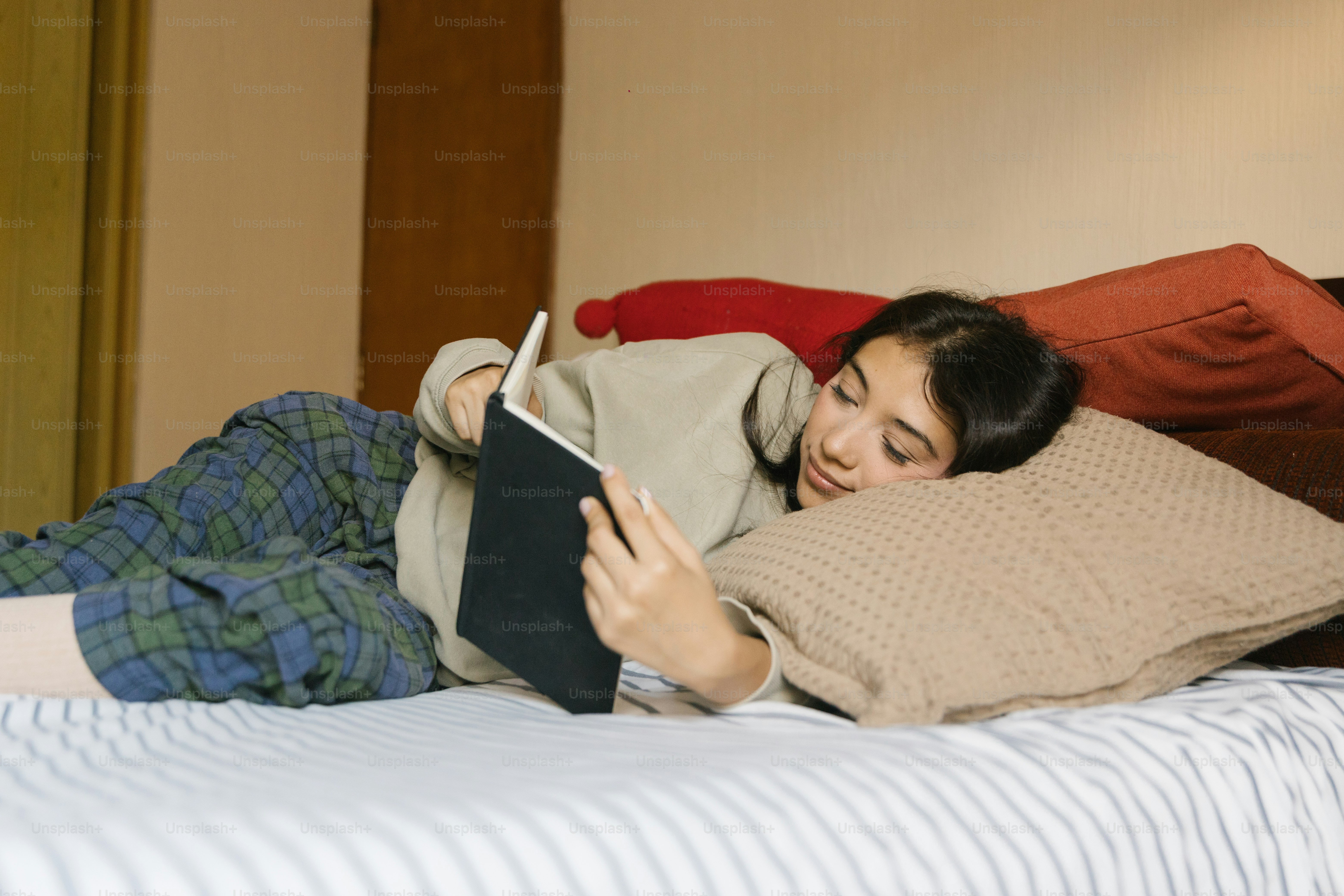 A person in a beige sweatshirt and plaid pants reading a book on a bed