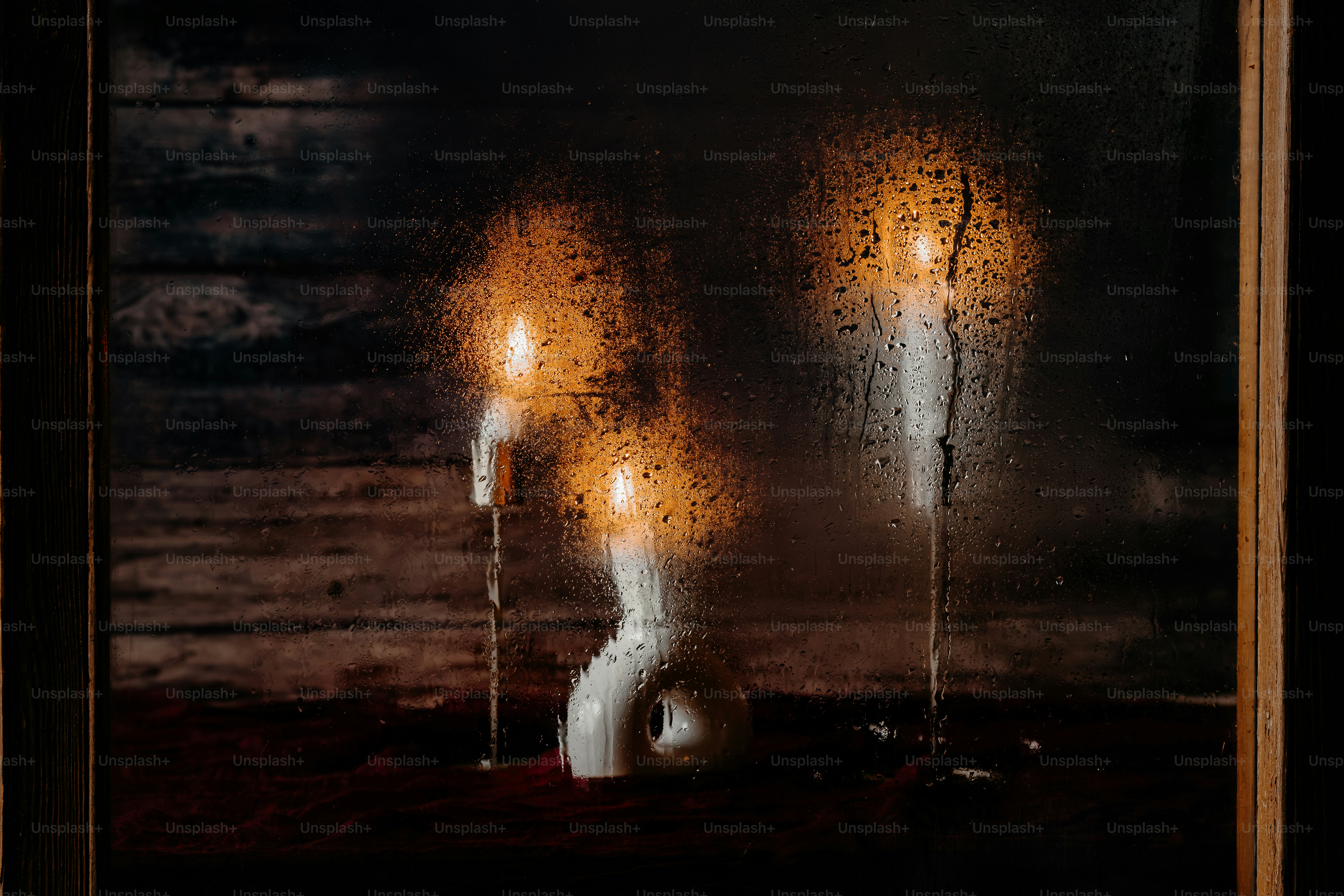 Three lit candles behind a rain-streaked window pane in a dark room