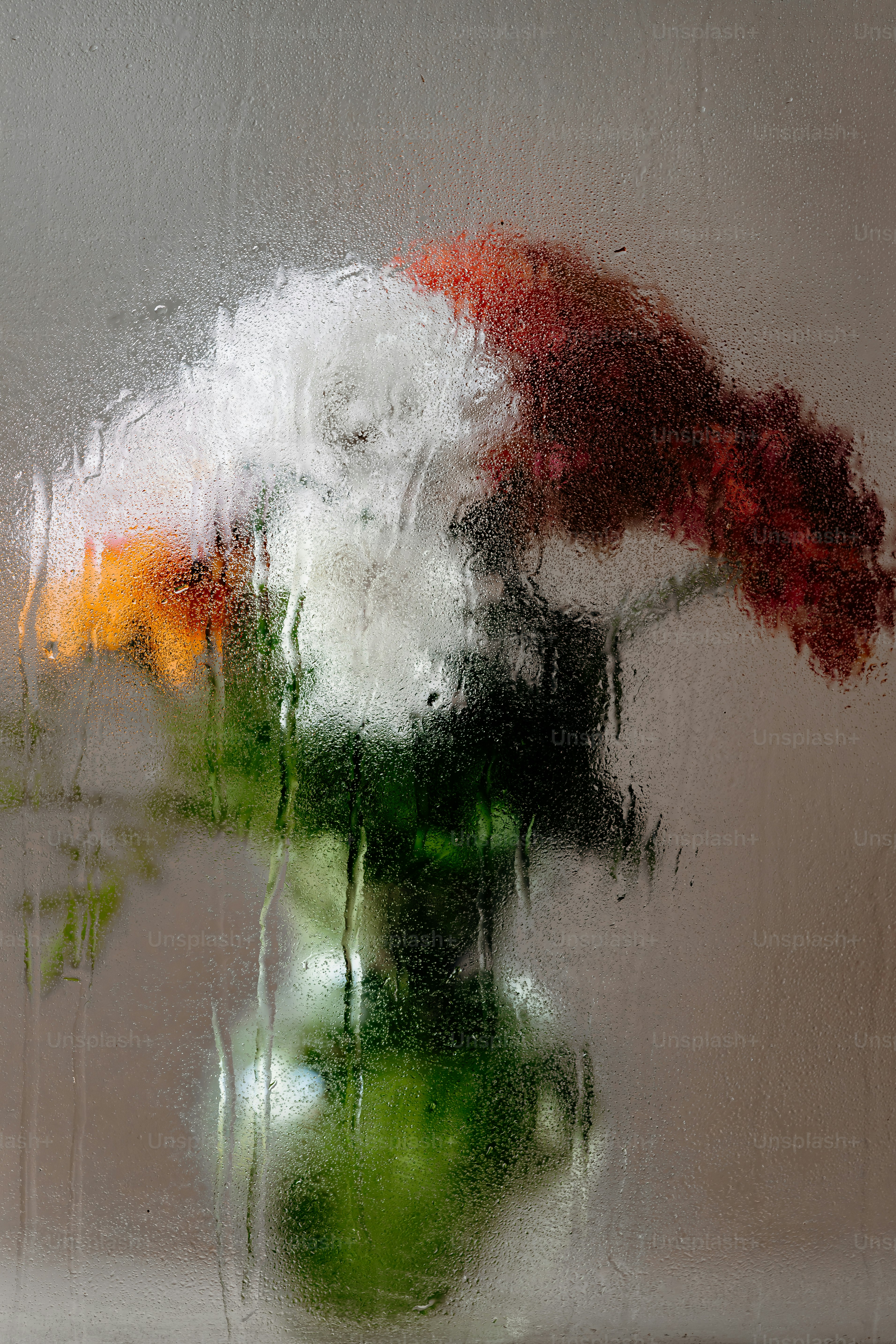A bouquet of white, orange, and red flowers behind a rain-streaked glass pane