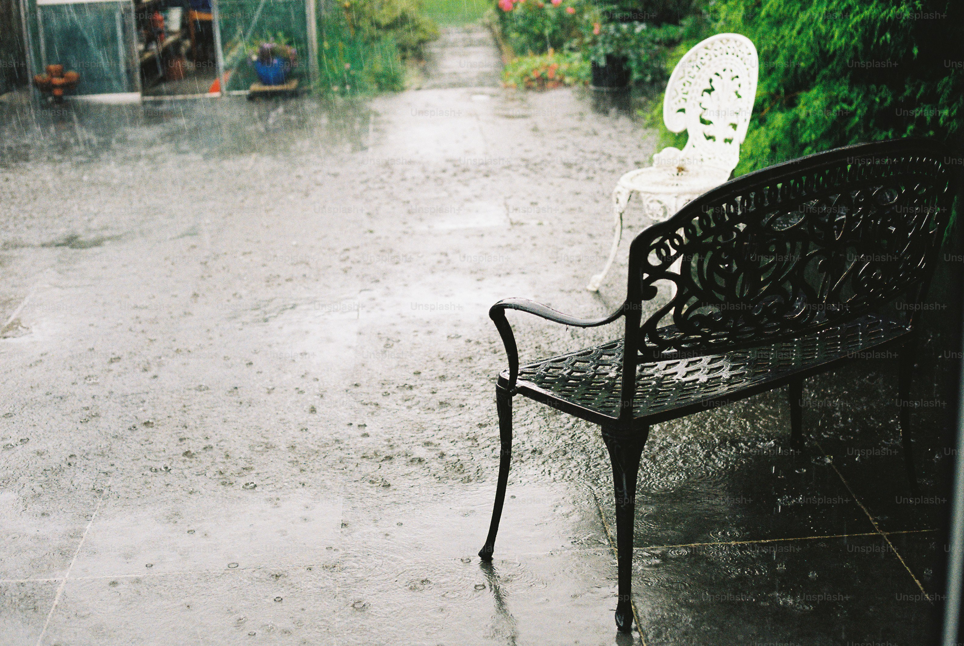 A black metal bench and a white chair on a wet patio during rain