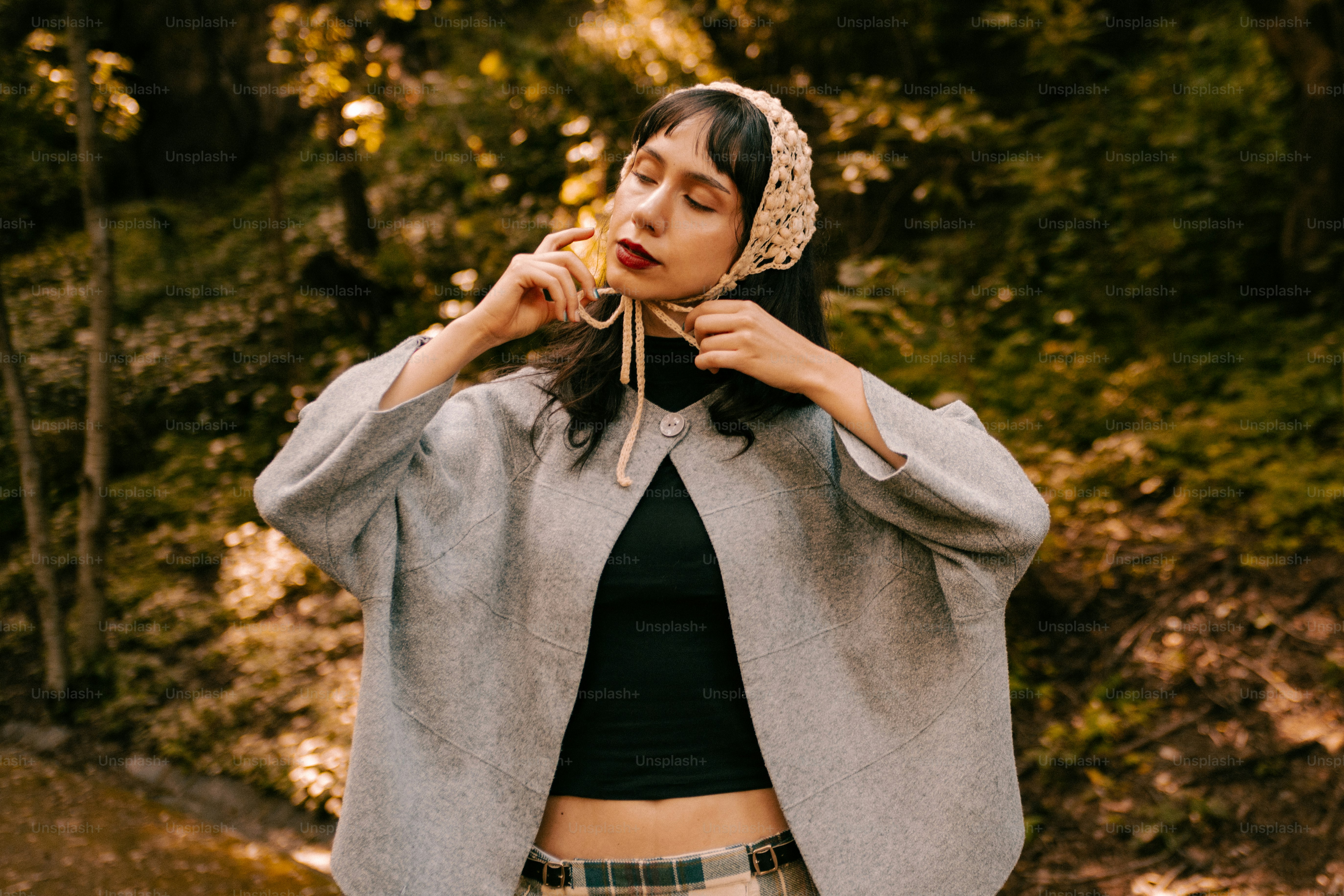 A woman in a grey coat and crochet headscarf in a sunlit forest
