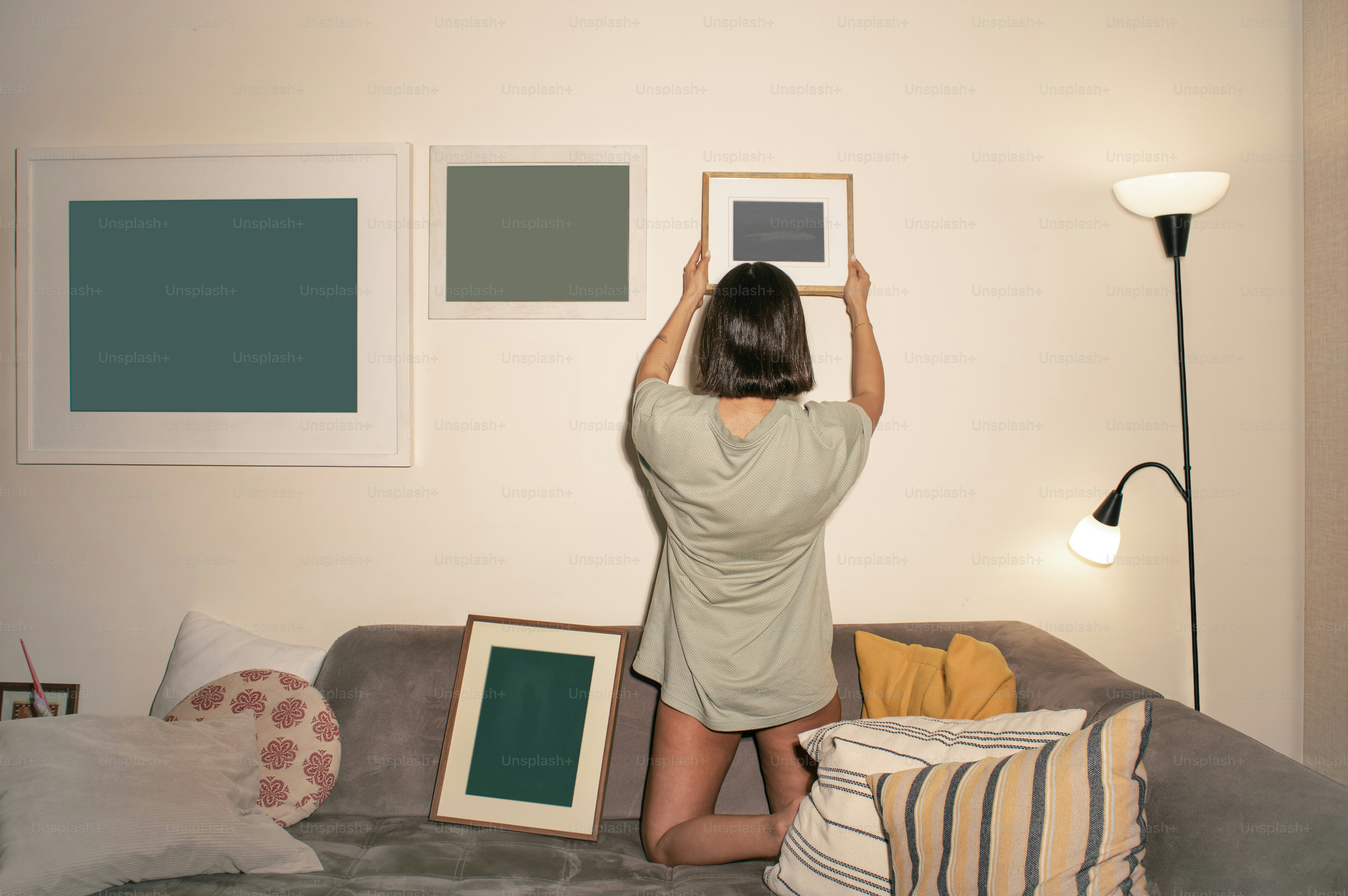 A woman kneeling on a grey sofa hanging a framed picture on a wall
