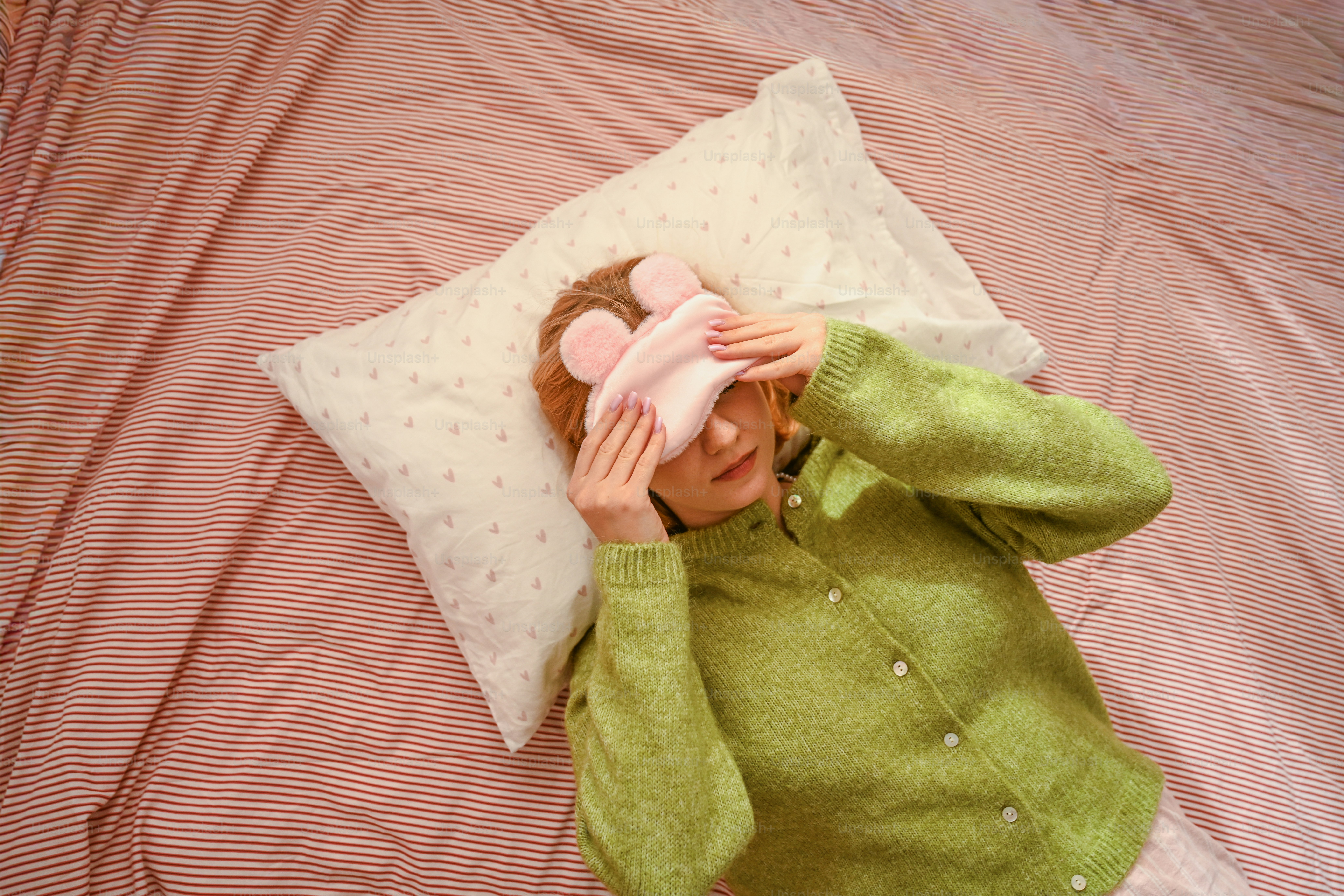 A woman in a green cardigan wearing a pink eye mask on a striped bed