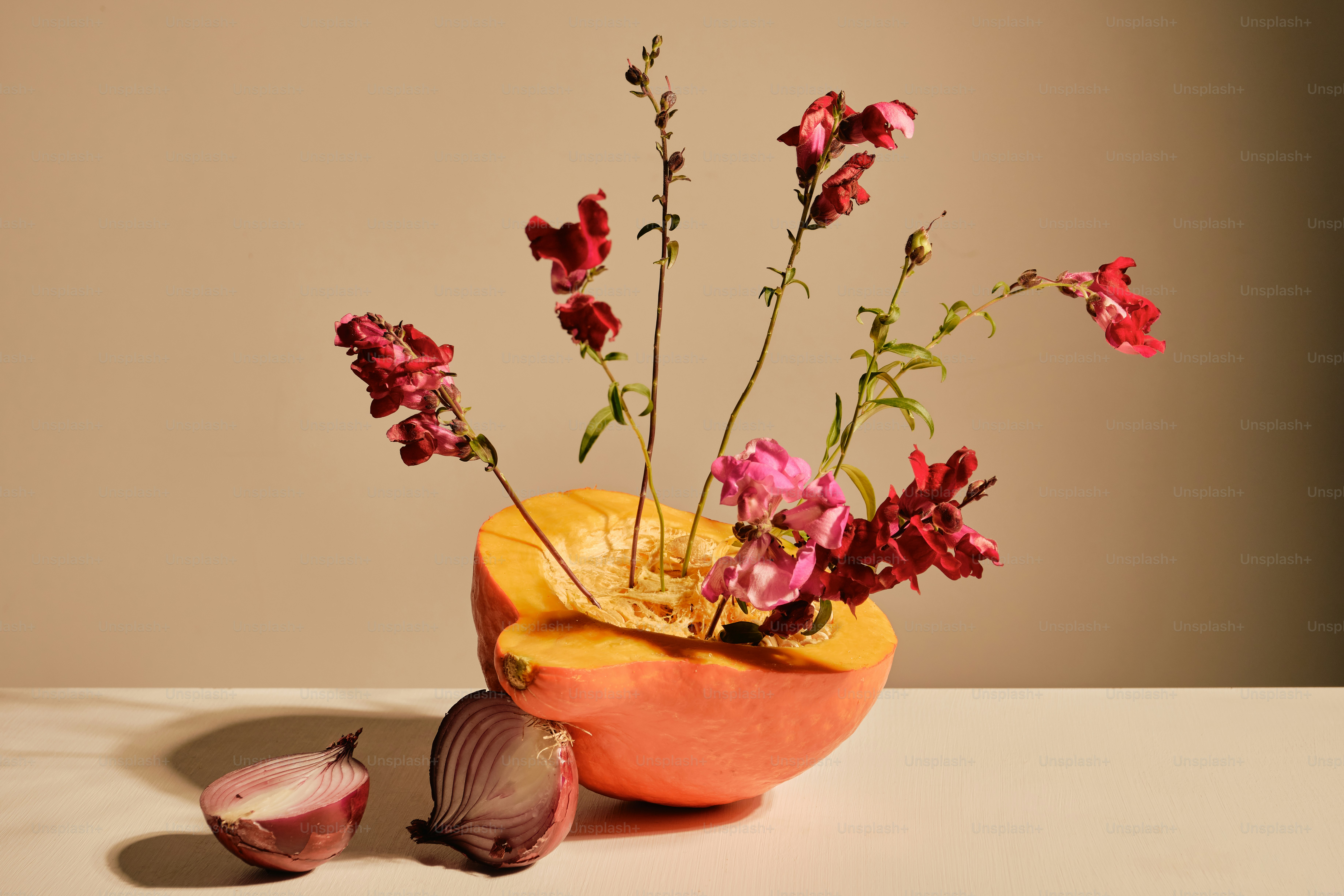 Red snapdragon flowers arranged in a halved pumpkin beside a halved red onion