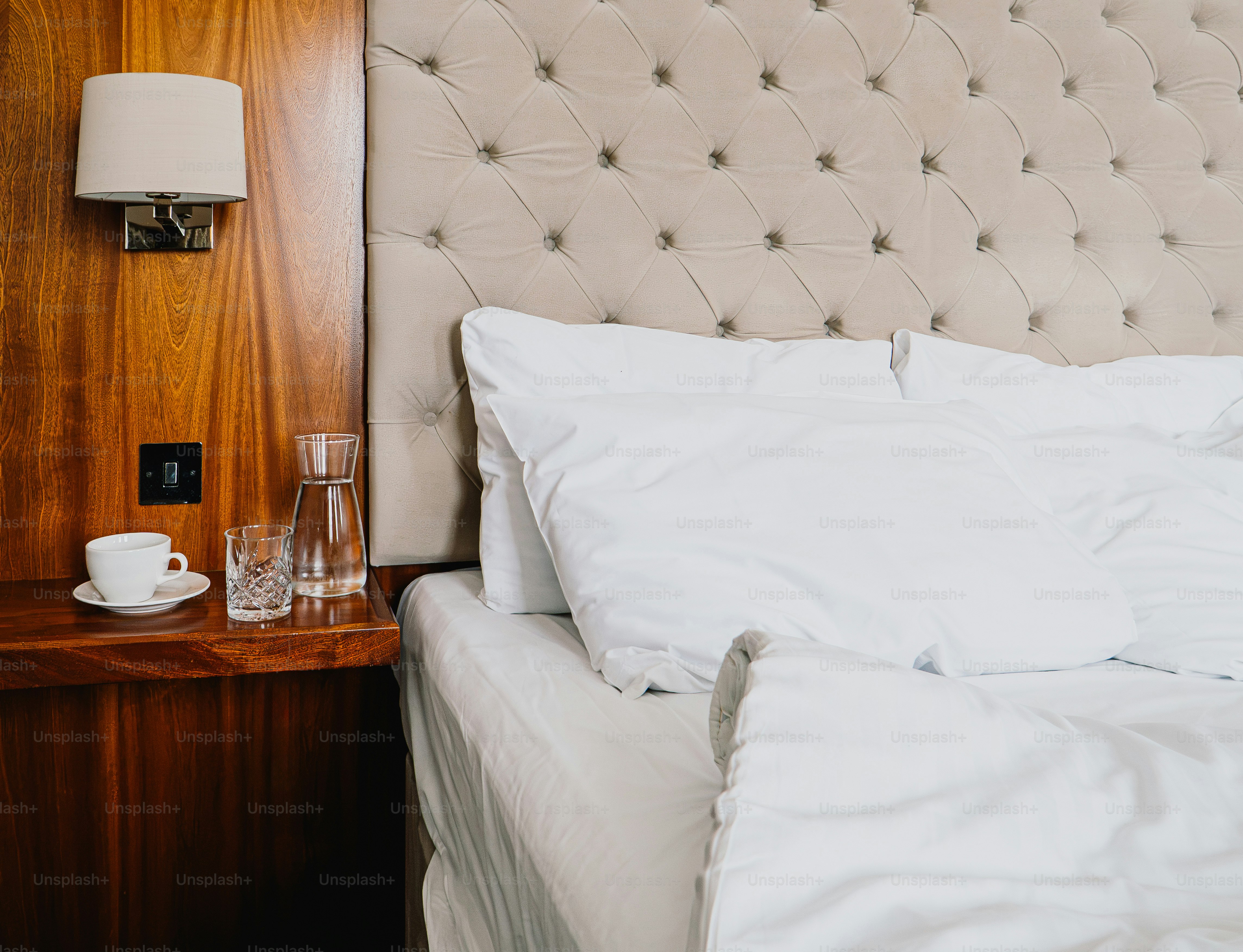 A white cup, glass, and water carafe on a wooden nightstand beside a bed