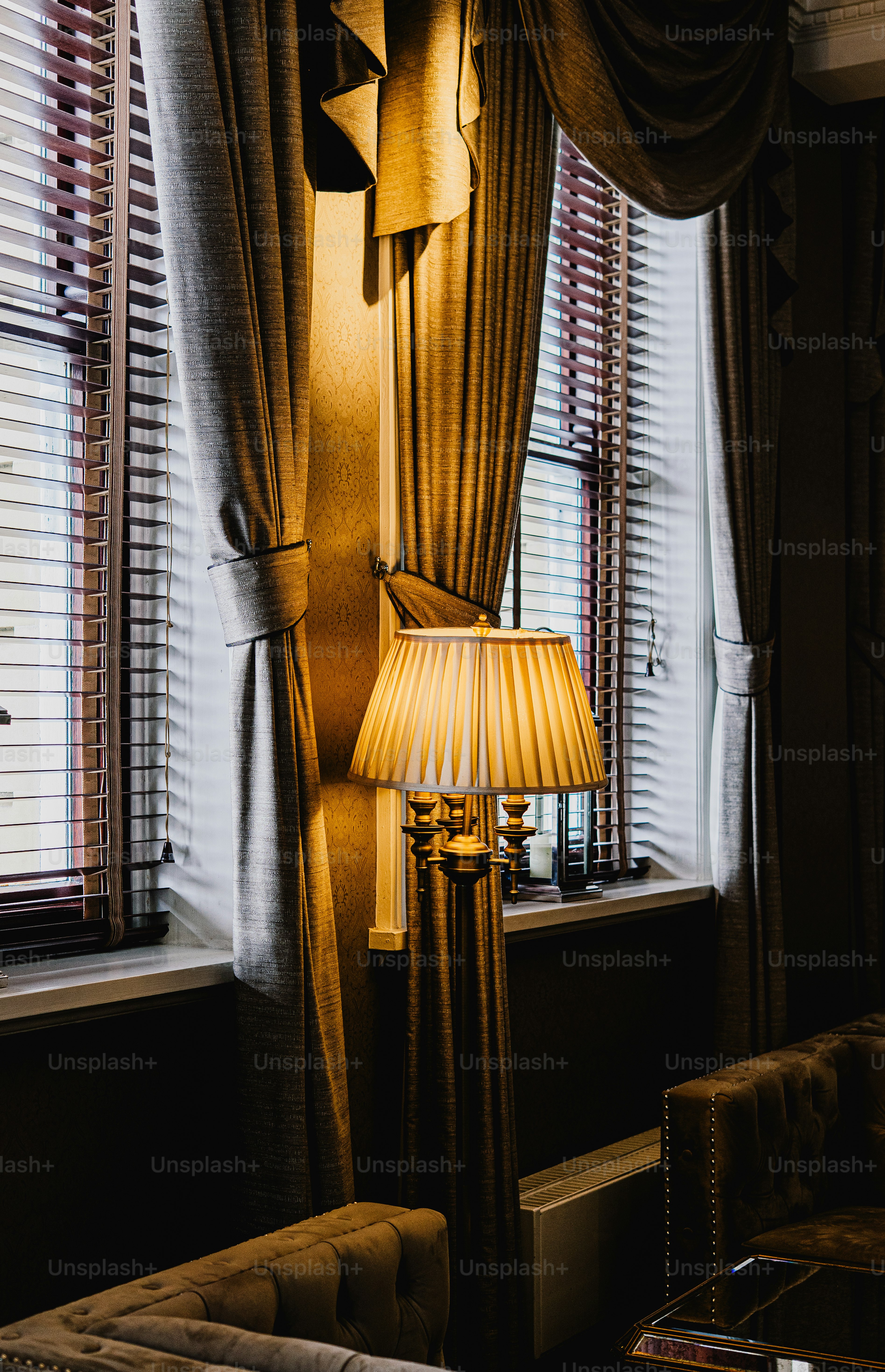 A floor lamp with a pleated shade glowing between tall windows and heavy drapes