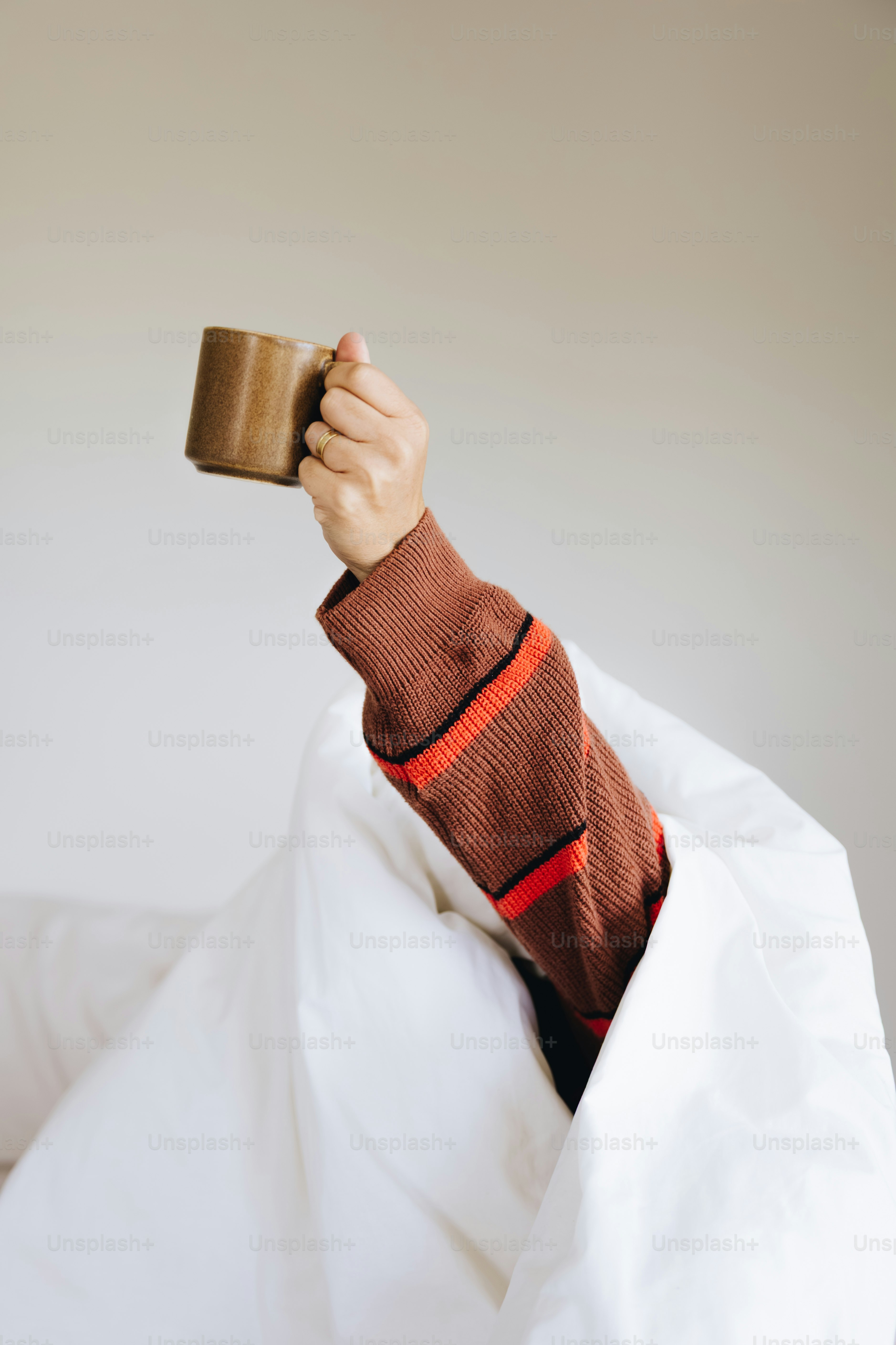 A person in a brown sweater holding a brown mug under white bedding