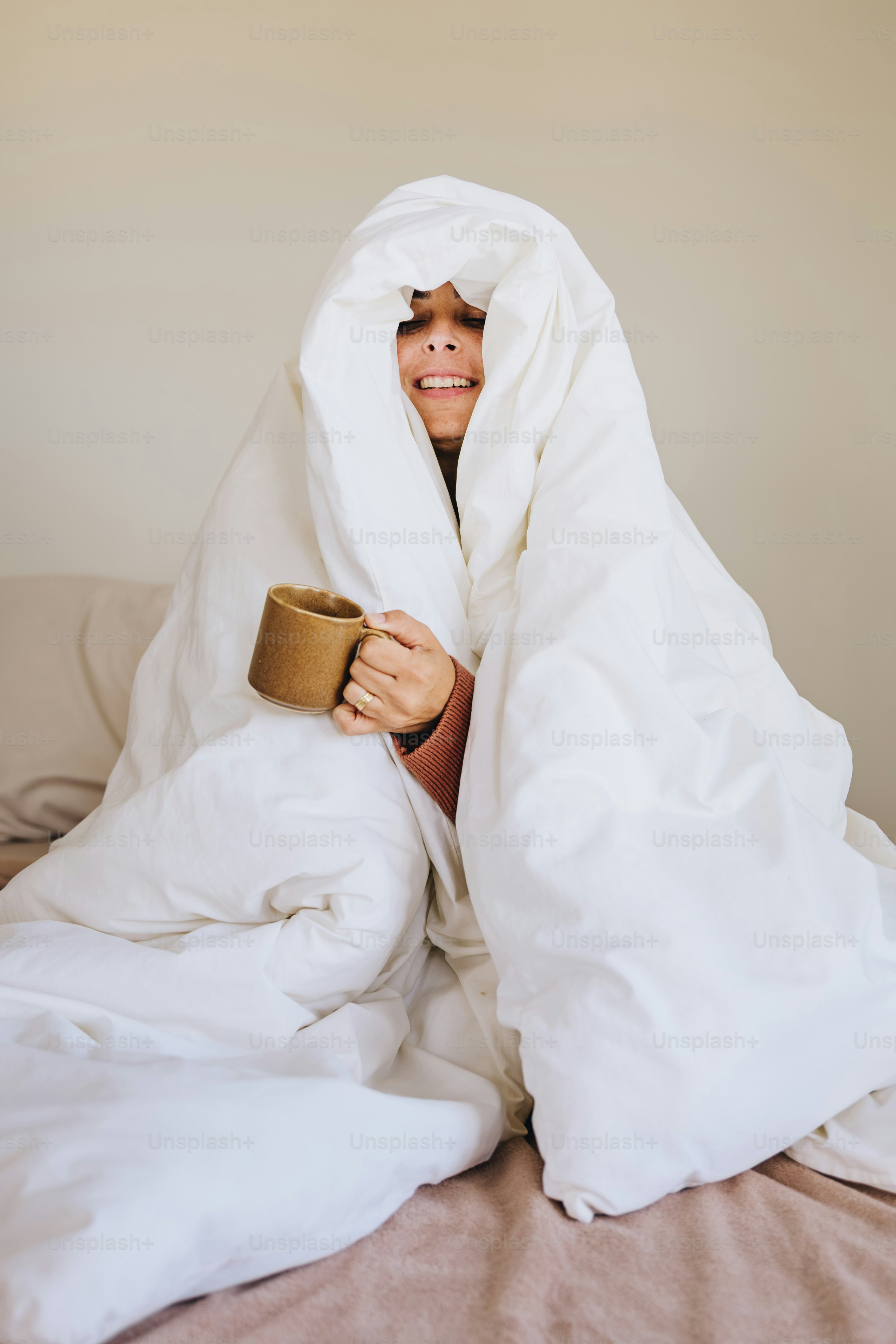 A smiling person wrapped in a white duvet holding a brown ceramic mug