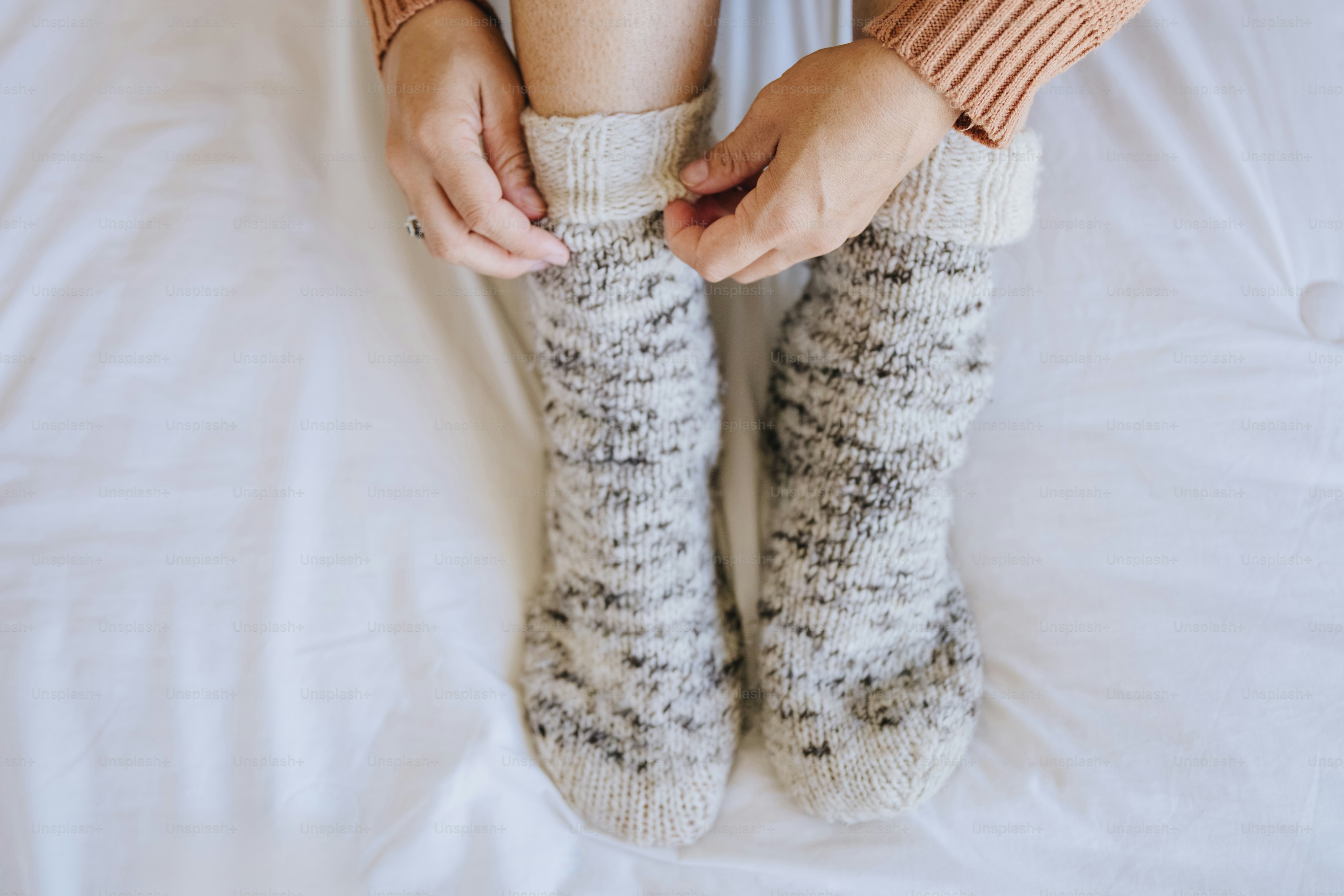 A person pulling on thick wool socks while sitting on a white bed