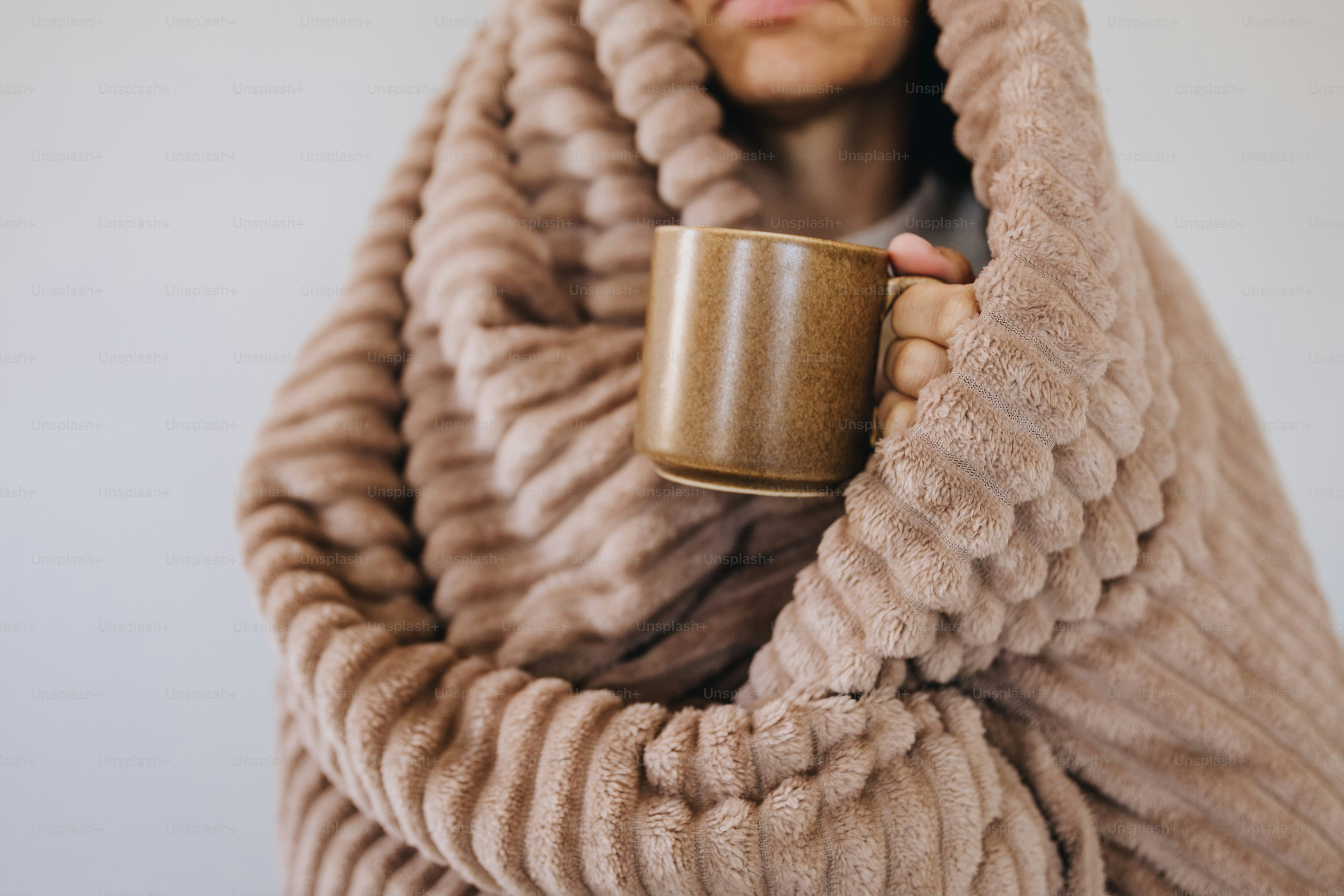 A person wrapped in a textured beige blanket holding a brown ceramic mug
