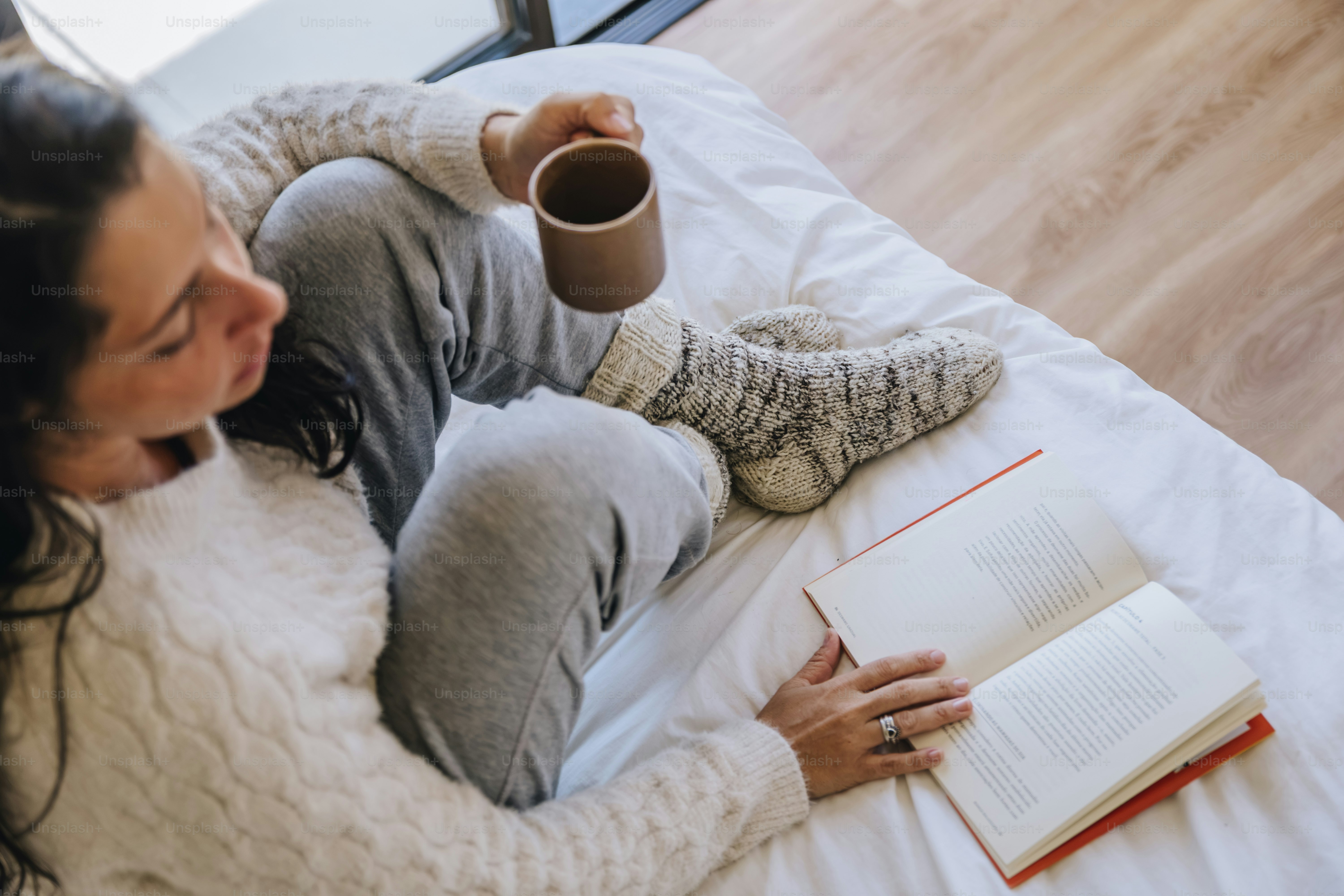 A woman in a cream sweater and grey sweatpants reading a book on a bed
